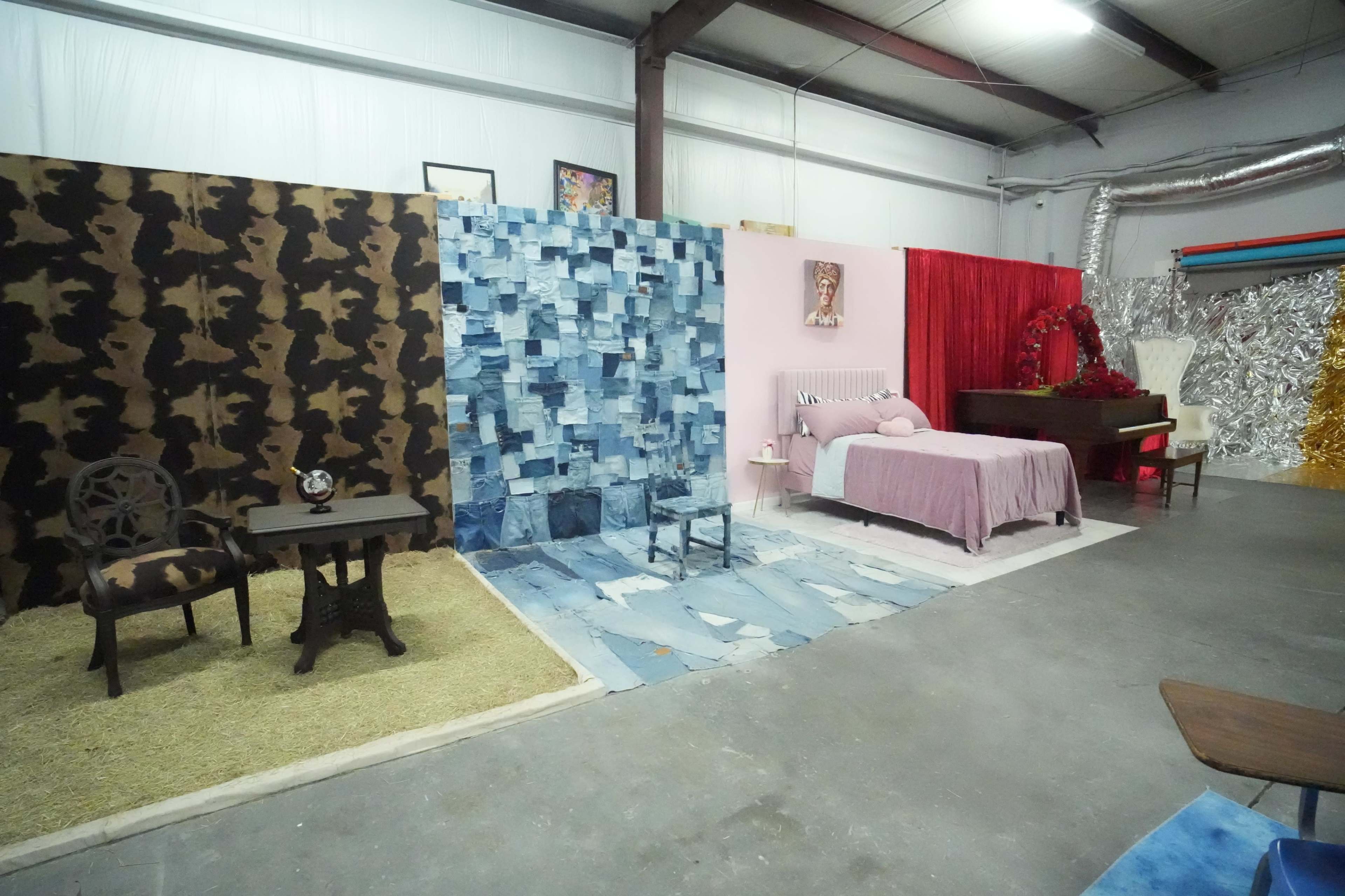 The image shows a large indoor space divided by colorful backdrops, featuring a bedroom setup with a bed, a piano, and various decorative elements.