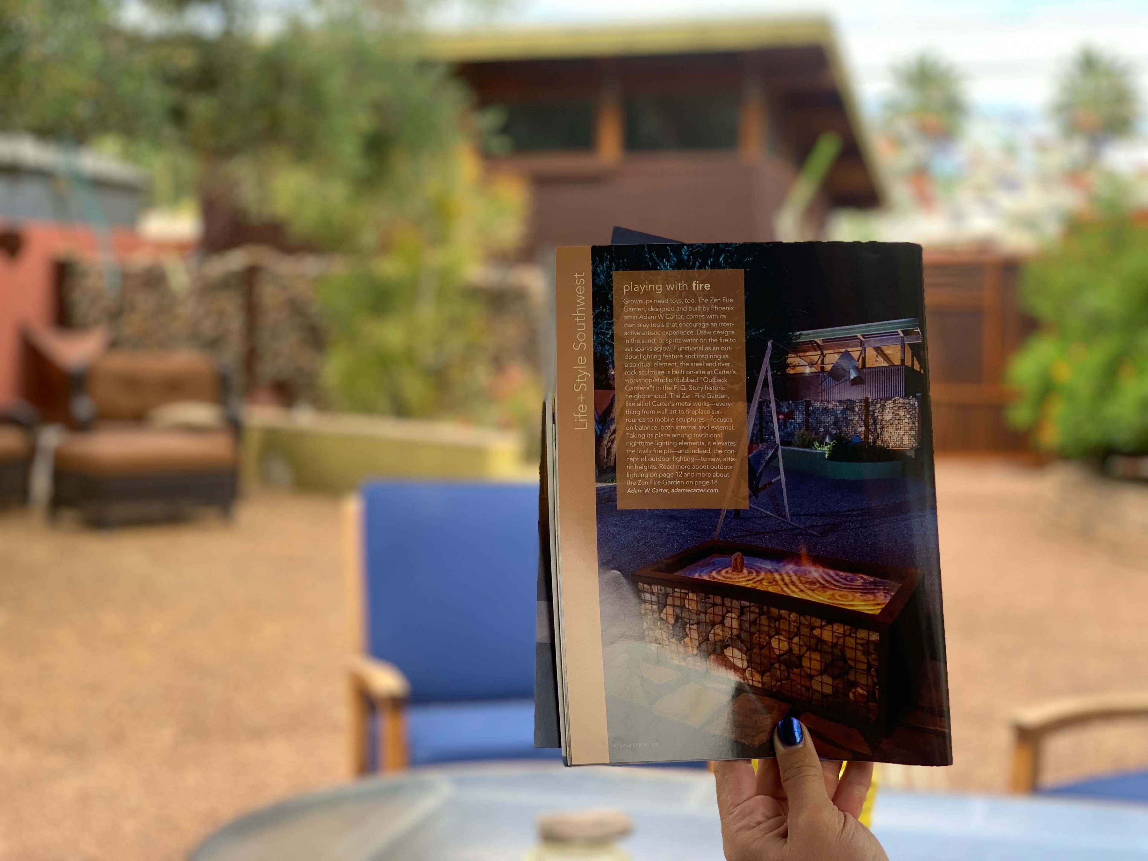 A person holds an open magazine featuring an article about a modern outdoor fire pit, with a backyard setting visible in the background.