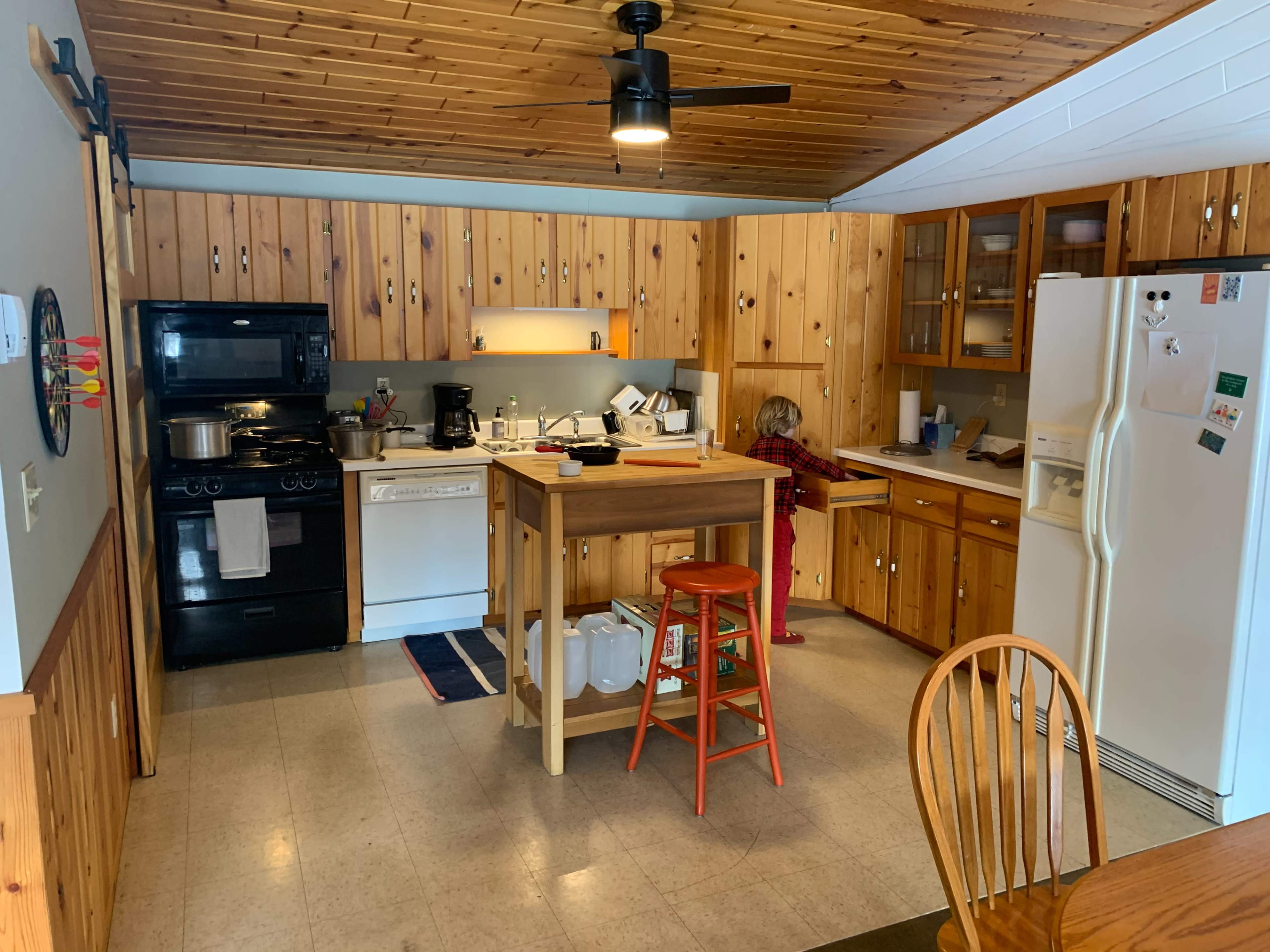 A kitchen features wooden cabinetry, a central island with a stool, and various appliances, including a stove, dishwasher, and refrigerator.