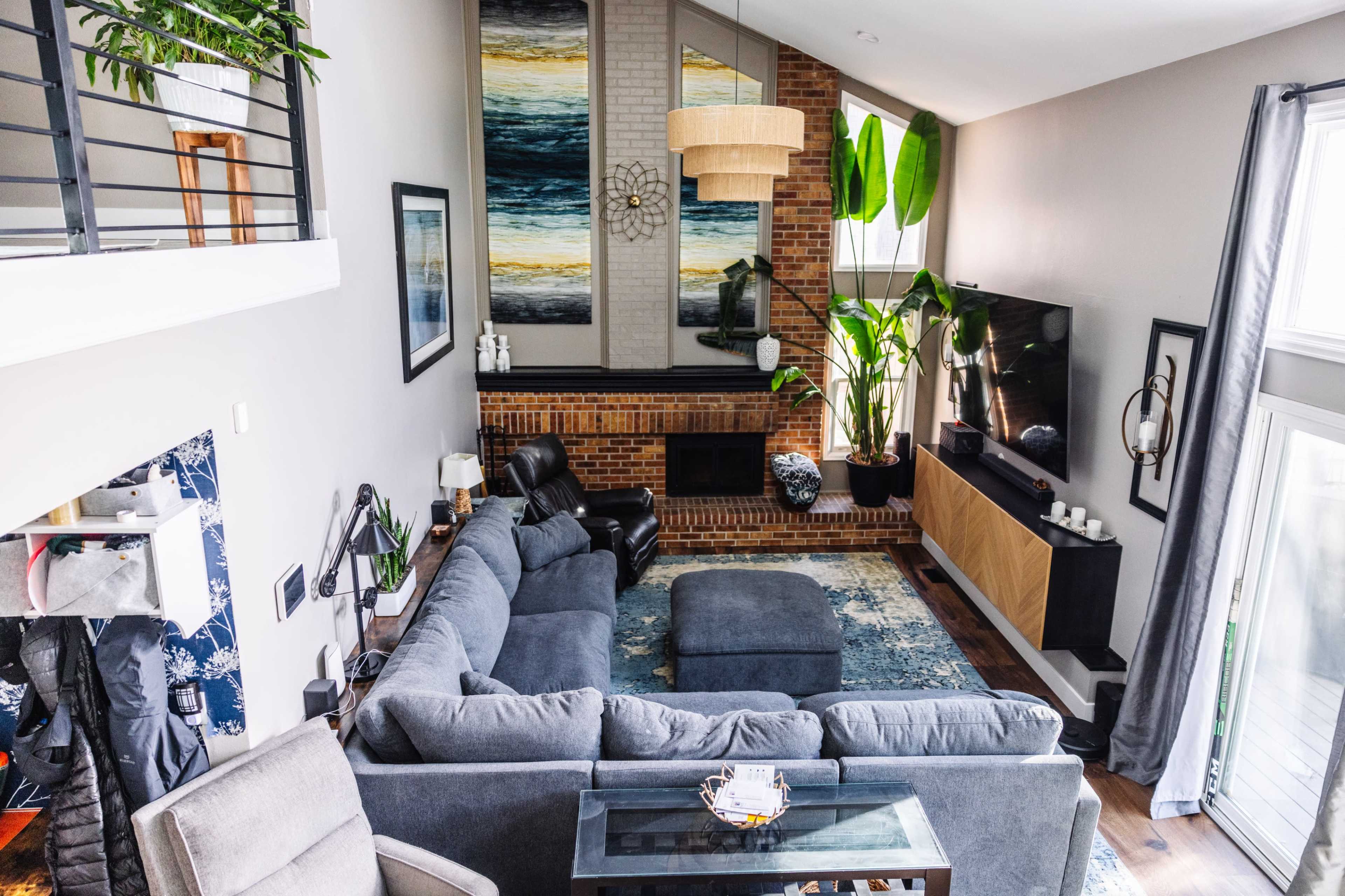 The image shows a modern living room with a large sectional sofa, a coffee table, a flat-screen TV mounted on the wall, and a tall plant near a brick fireplace.
