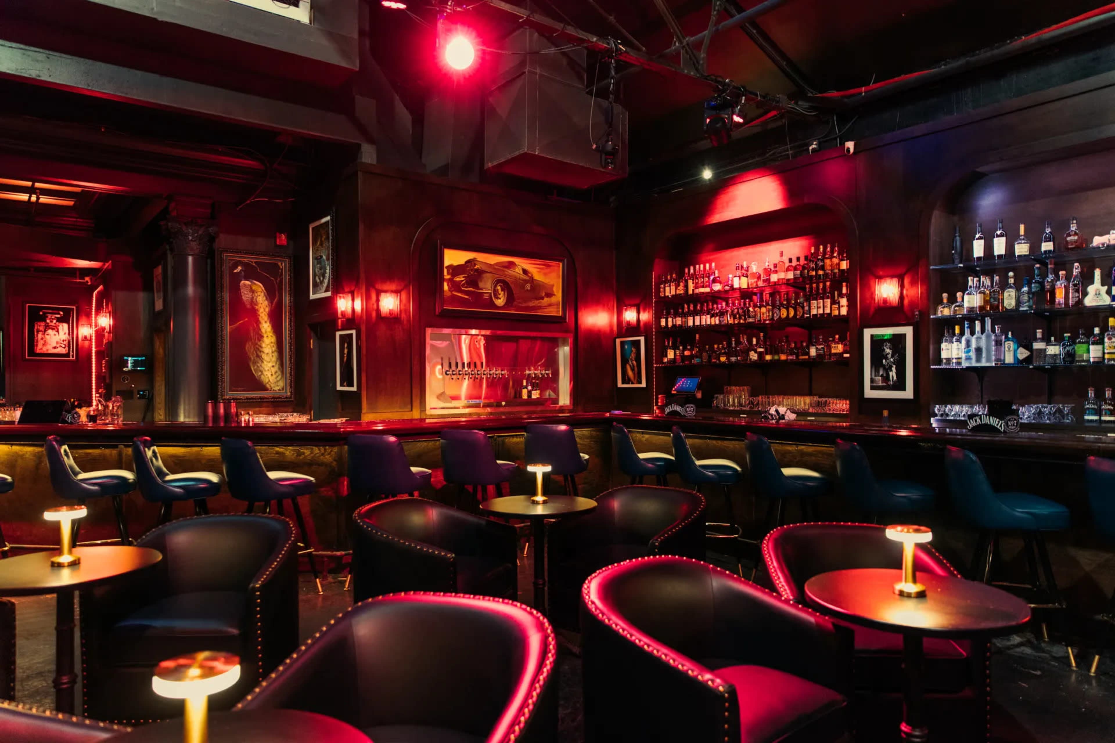 The image shows a dimly lit bar featuring dark wood paneling, a well-stocked backlit bar shelf, and seating arranged with small tables and plush chairs.