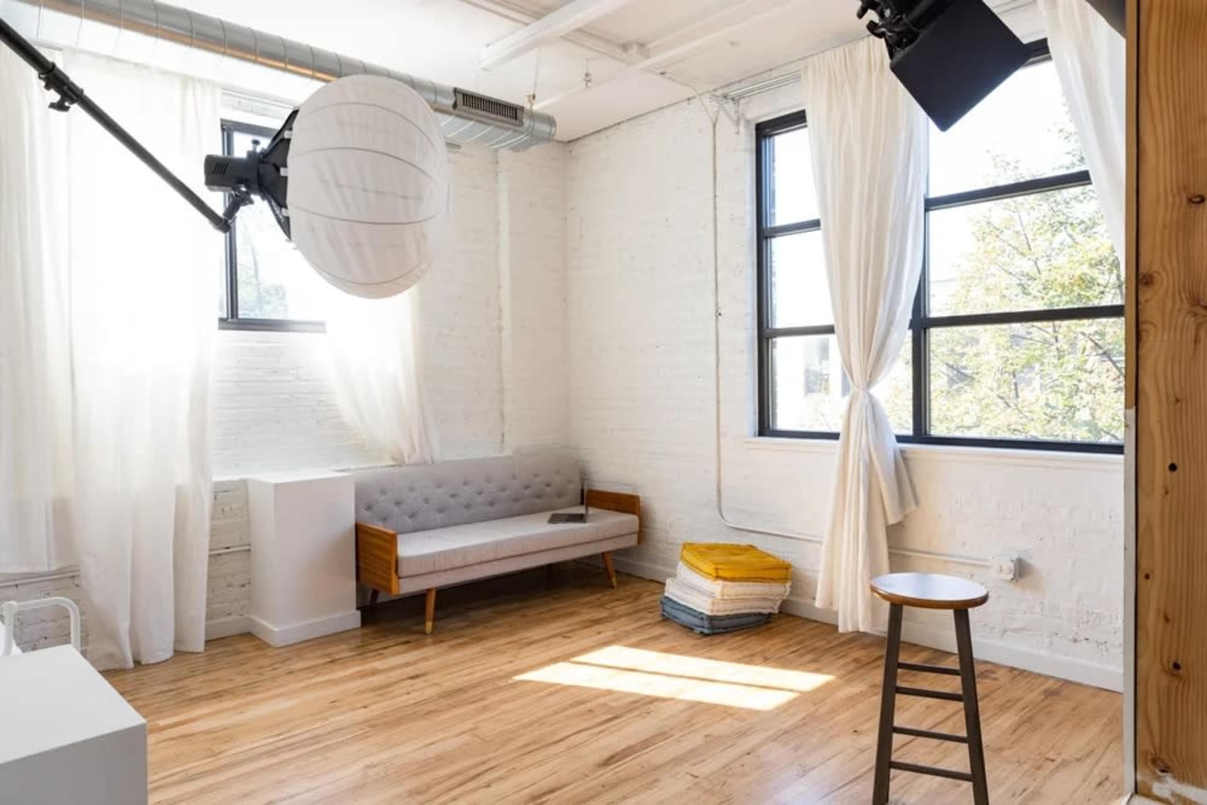 The image shows a bright, well-lit room featuring a gray sofa, a wooden stool, and large windows with sheer white curtains.