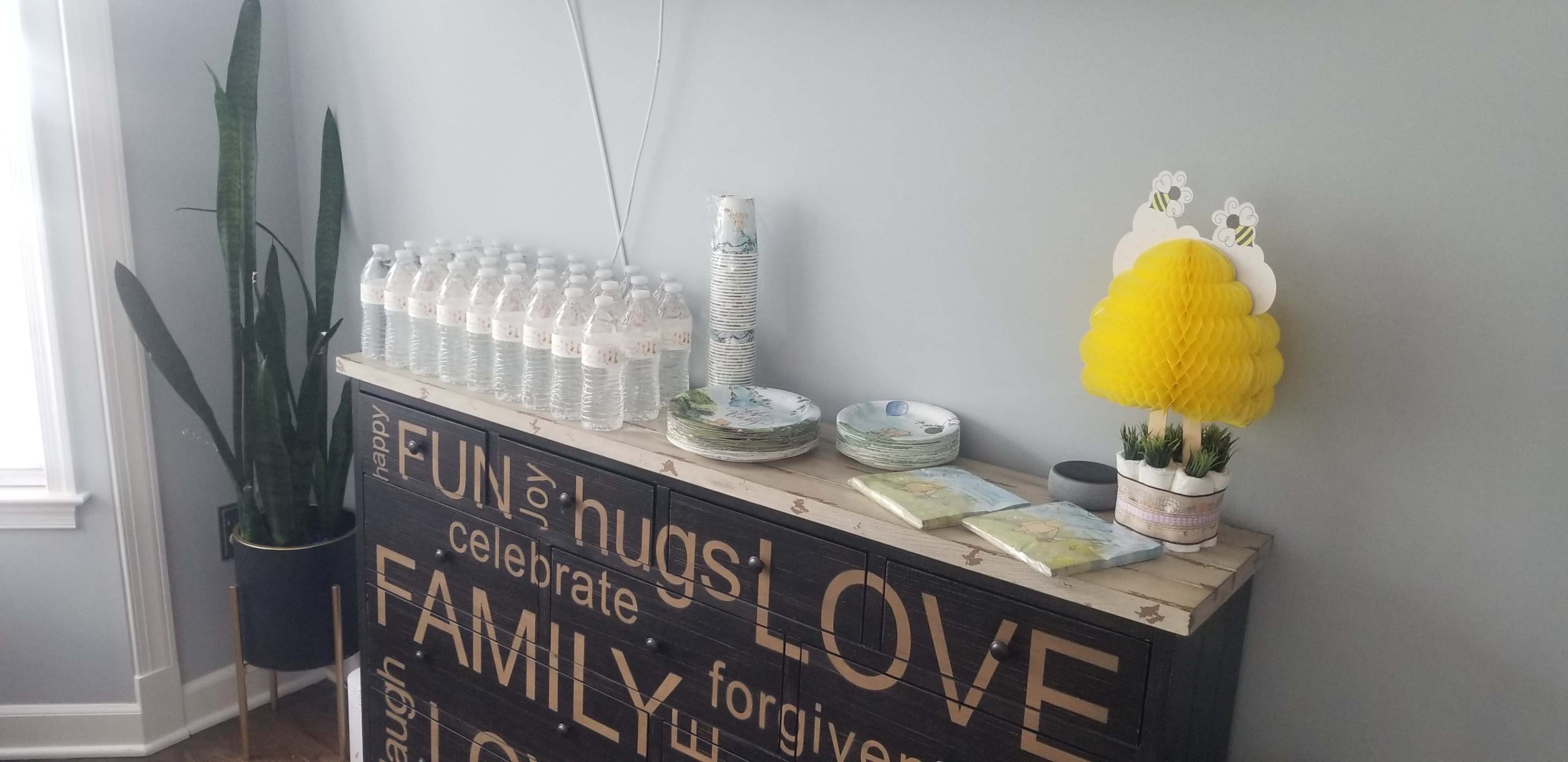 A buffet table decorated with bottled water, disposable cups, plates, and a yellow decorative item is set against a light gray wall.