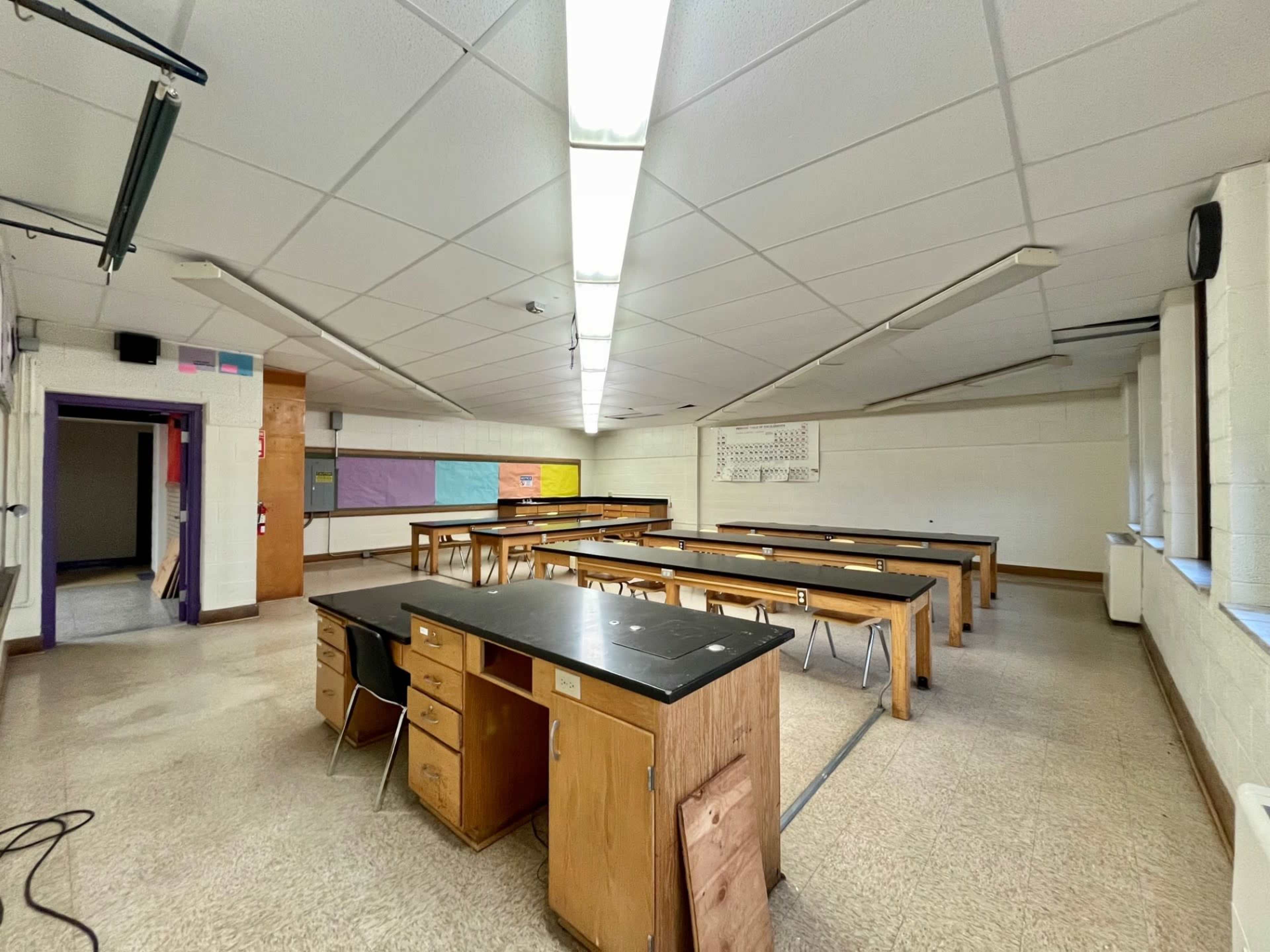 The image shows a spacious classroom with several long tables, chairs, and a large window, illuminated by fluorescent lights.