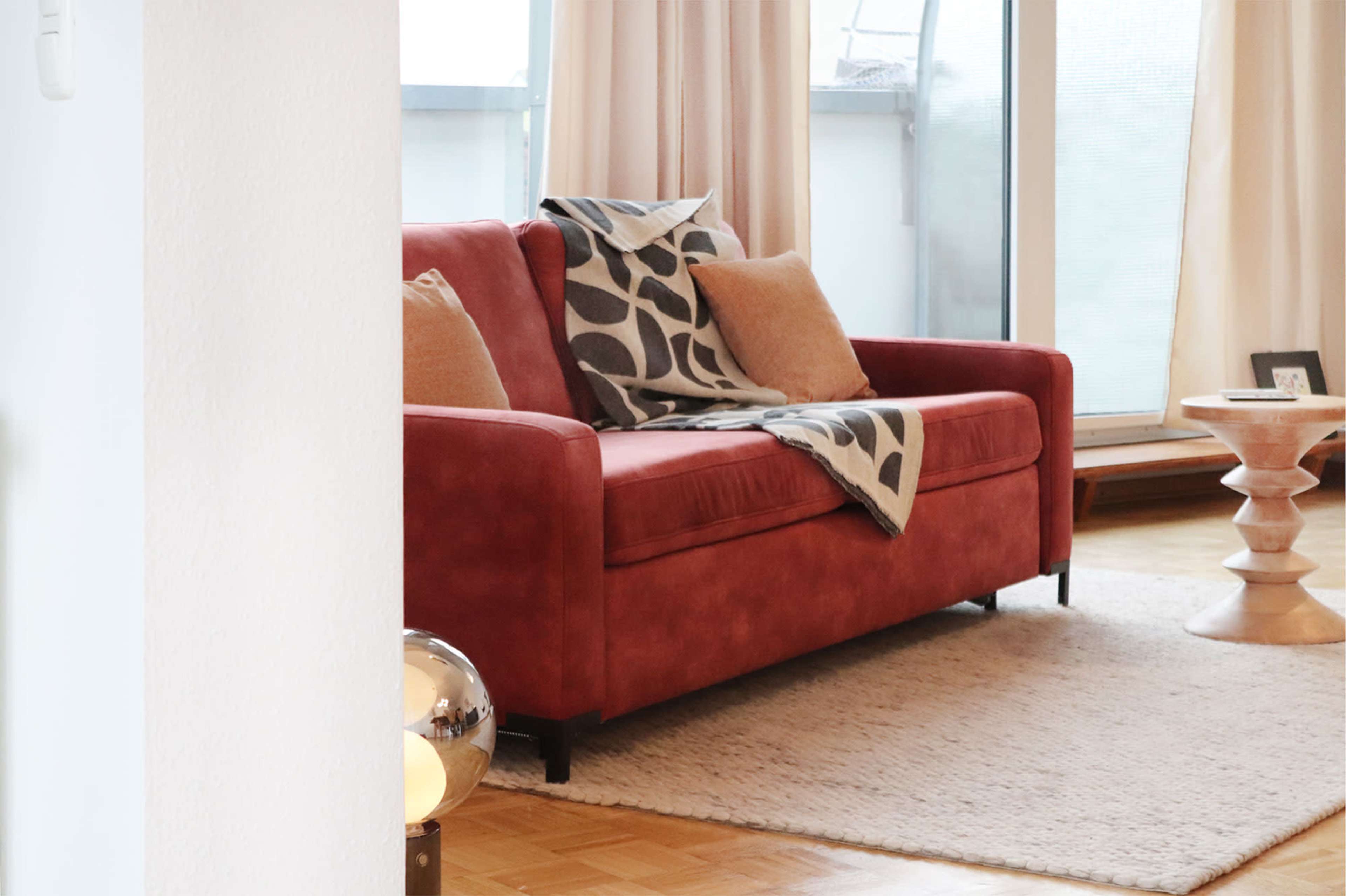 A red couch with pillows and a patterned throw is positioned in a bright living room near a textured rug and a wooden side table.