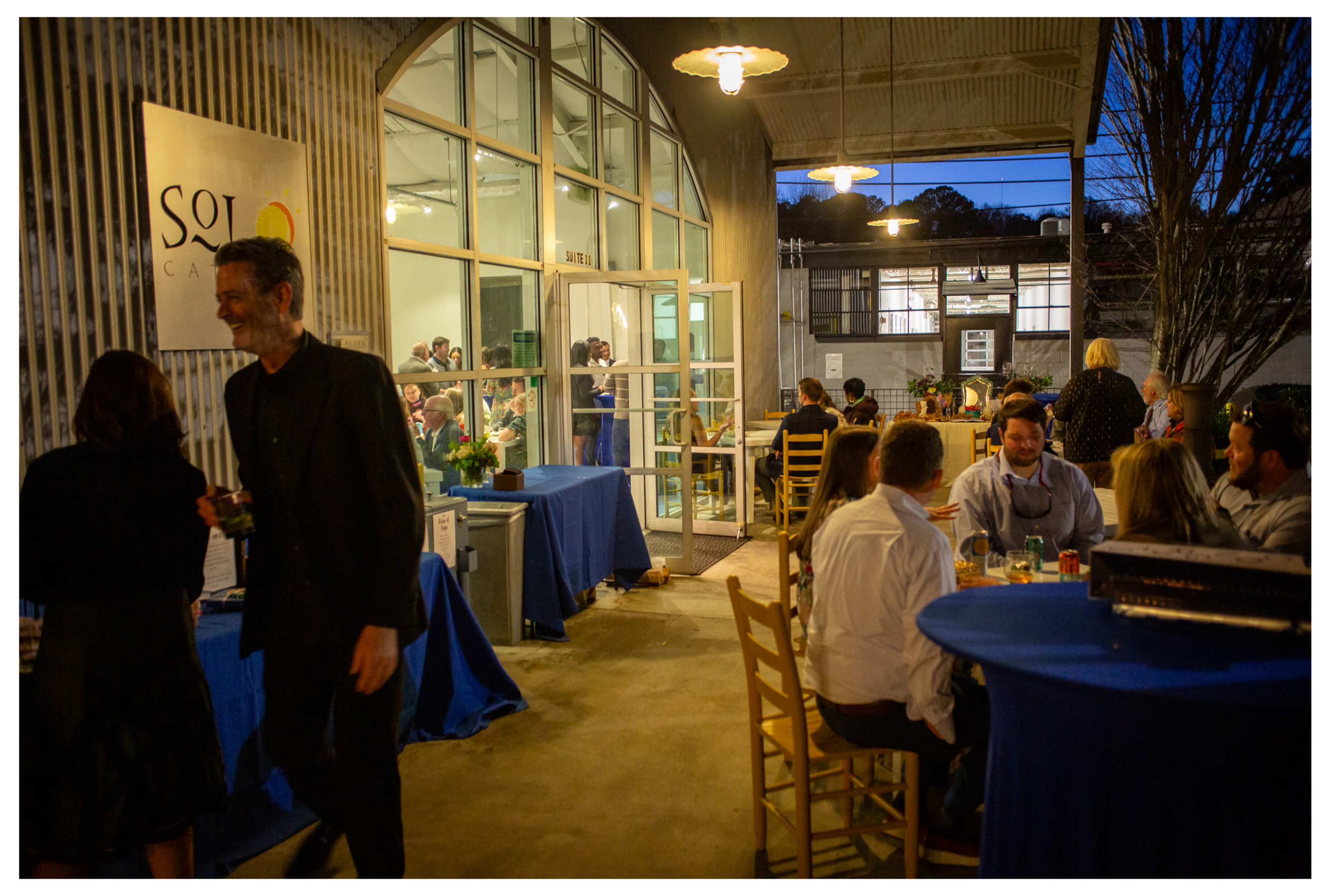 The image depicts a gathering inside a venue with tables set for dining, where people are mingling and enjoying drinks as dusk approaches outside.