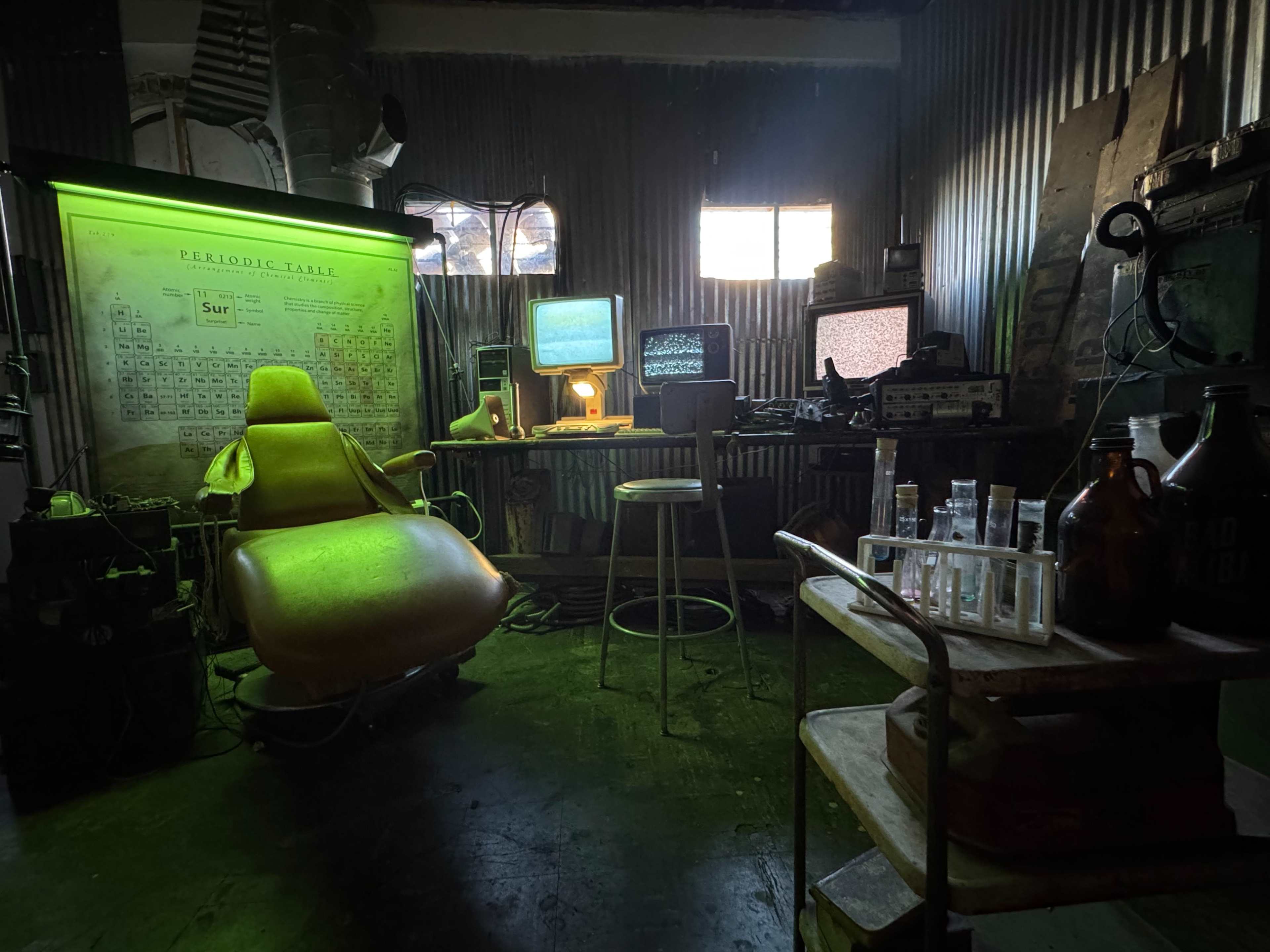 The image shows a dimly lit laboratory-like room with a vintage chair, multiple old computer monitors, a periodic table on the wall, and various lab equipment on a cart.