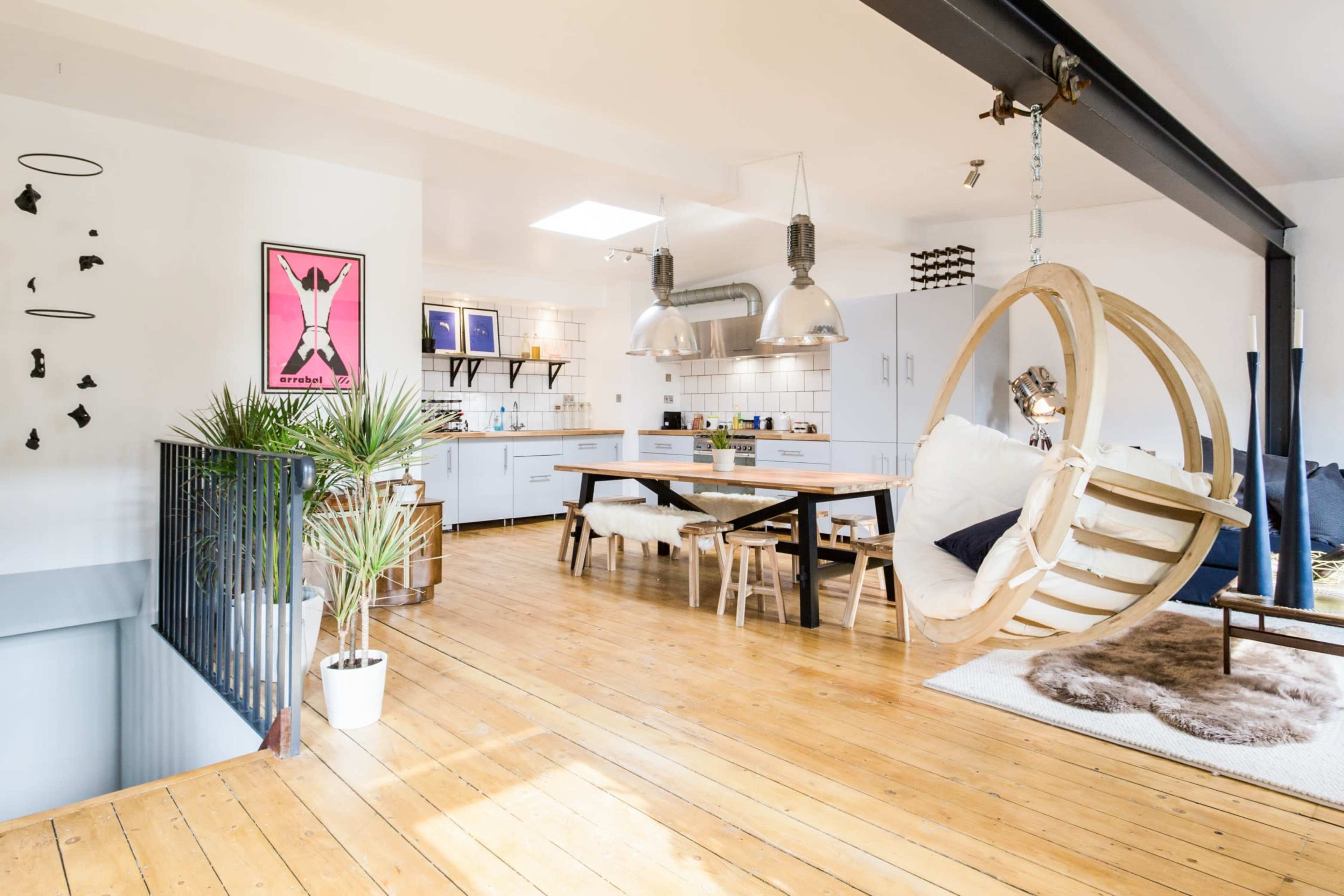 The image shows a modern open-plan kitchen and dining area with wooden floors, a long table surrounded by chairs, and a hanging chair in the corner.