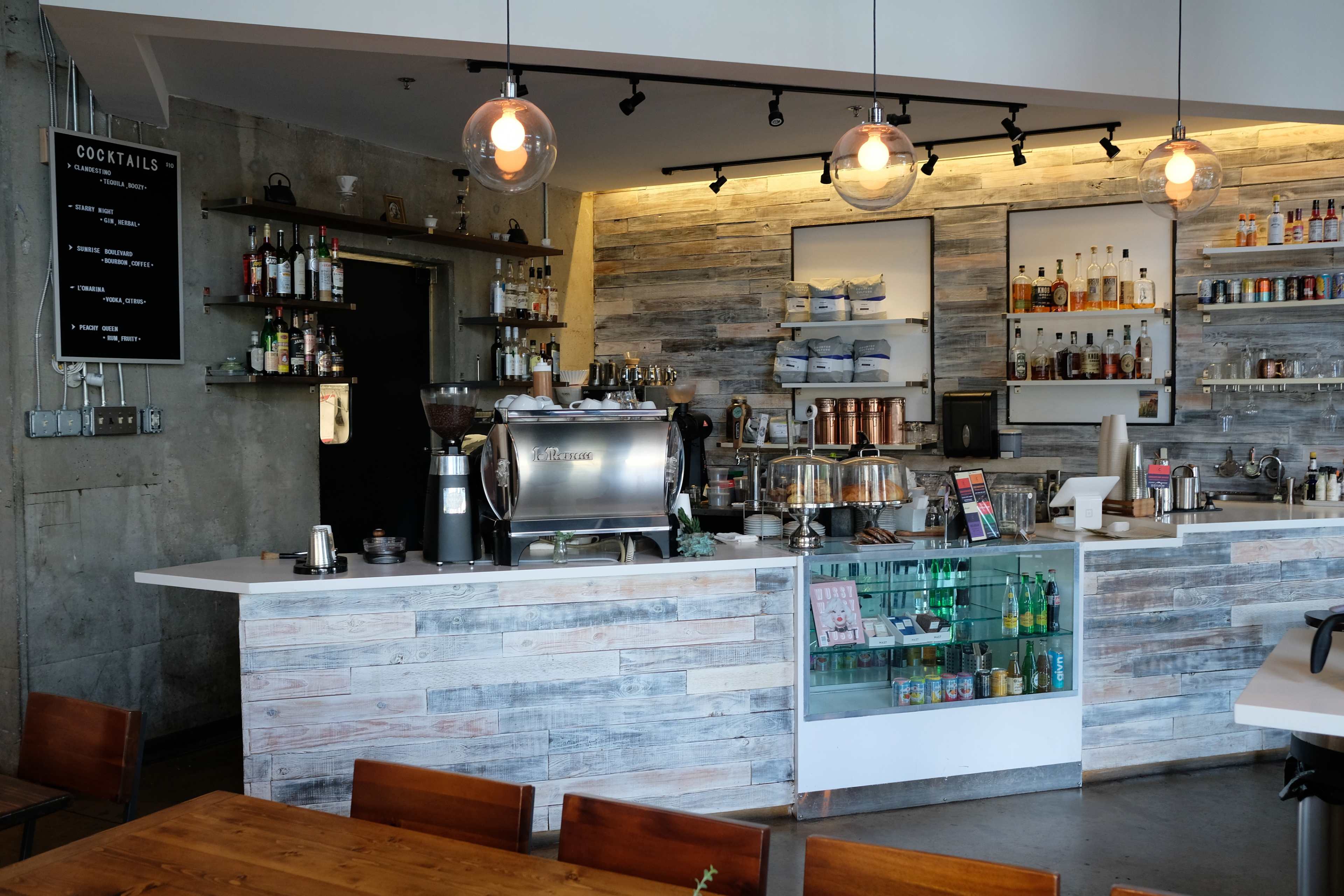 A coffee shop interior features a wooden counter with an espresso machine, shelves of drinks, and pendant lighting.