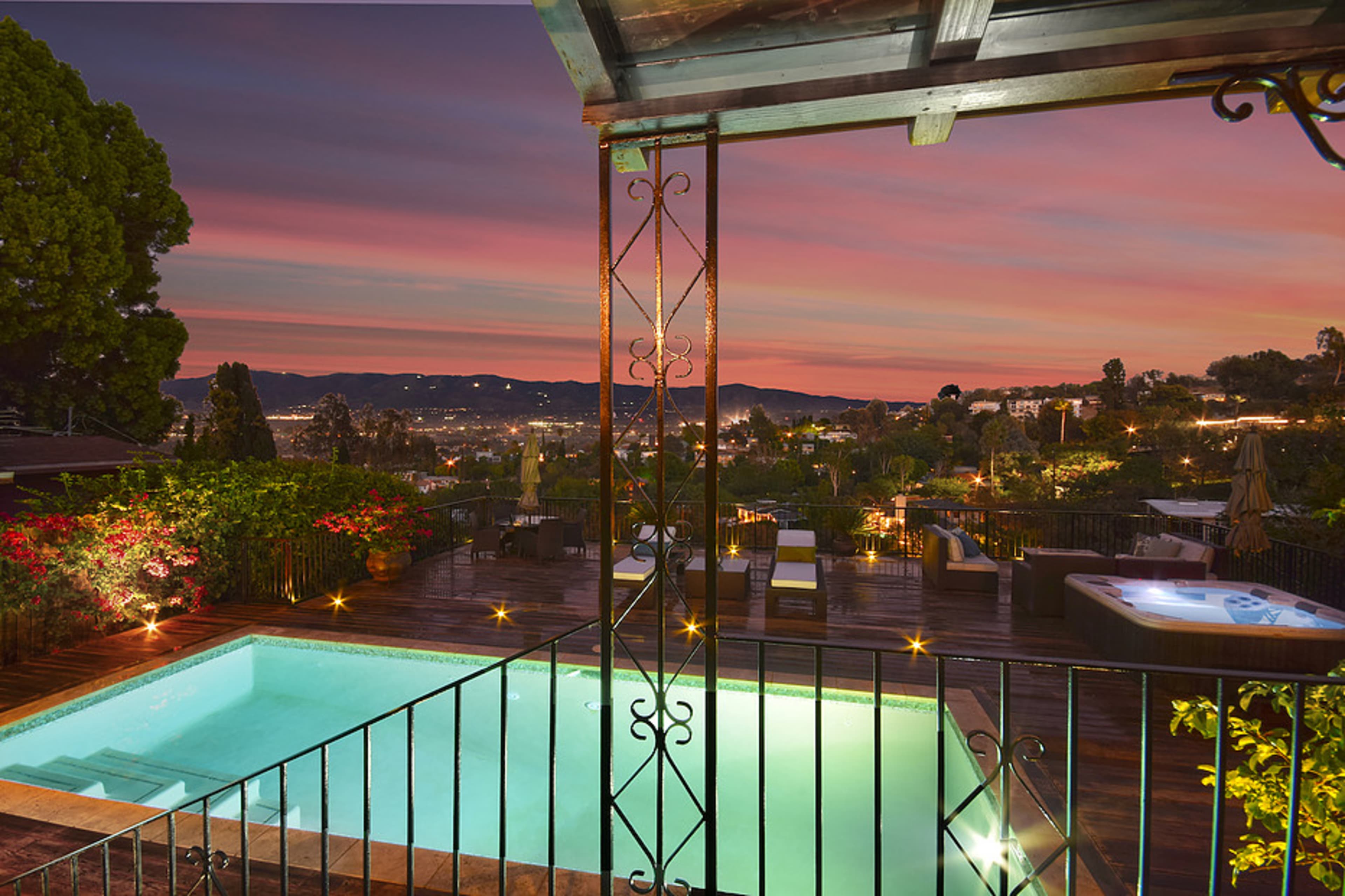 A swimming pool with a wooden deck overlooks a scenic landscape at sunset, surrounded by greenery and lit outdoor seating areas.