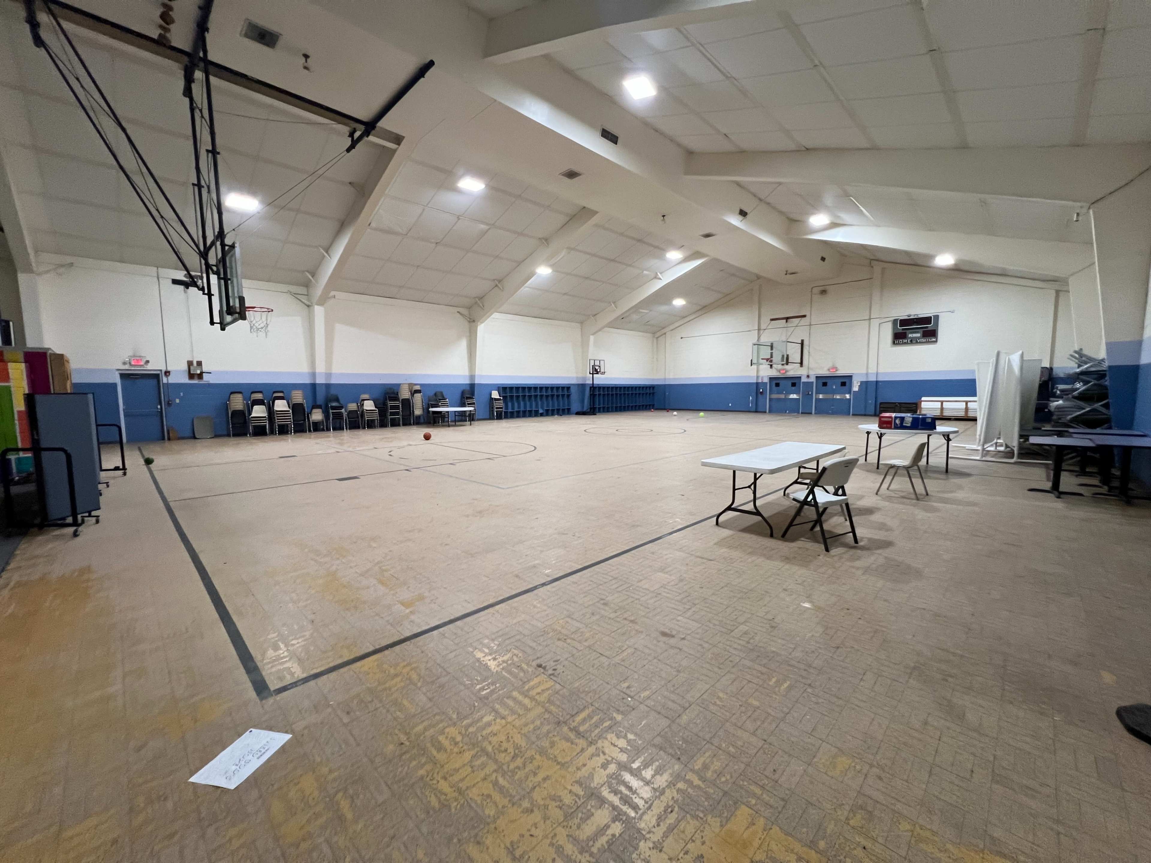The image shows an empty gymnasium with basketball hoops, chairs arranged along the wall, and a few tables set up on one side.