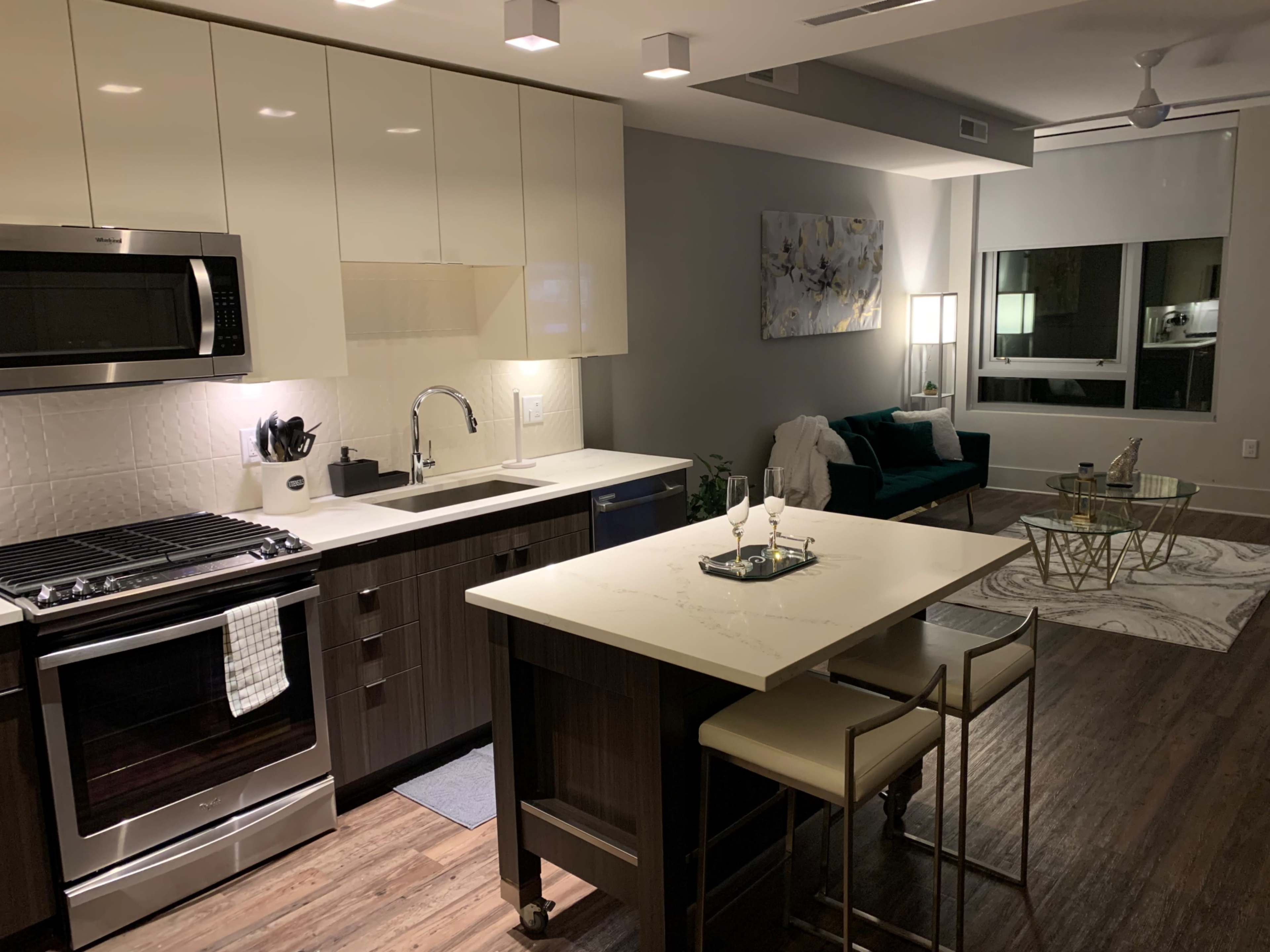 A modern kitchen with a stainless steel refrigerator, an oven, and a central island with two bar stools, adjacent to a living space featuring a green couch and a coffee table.
