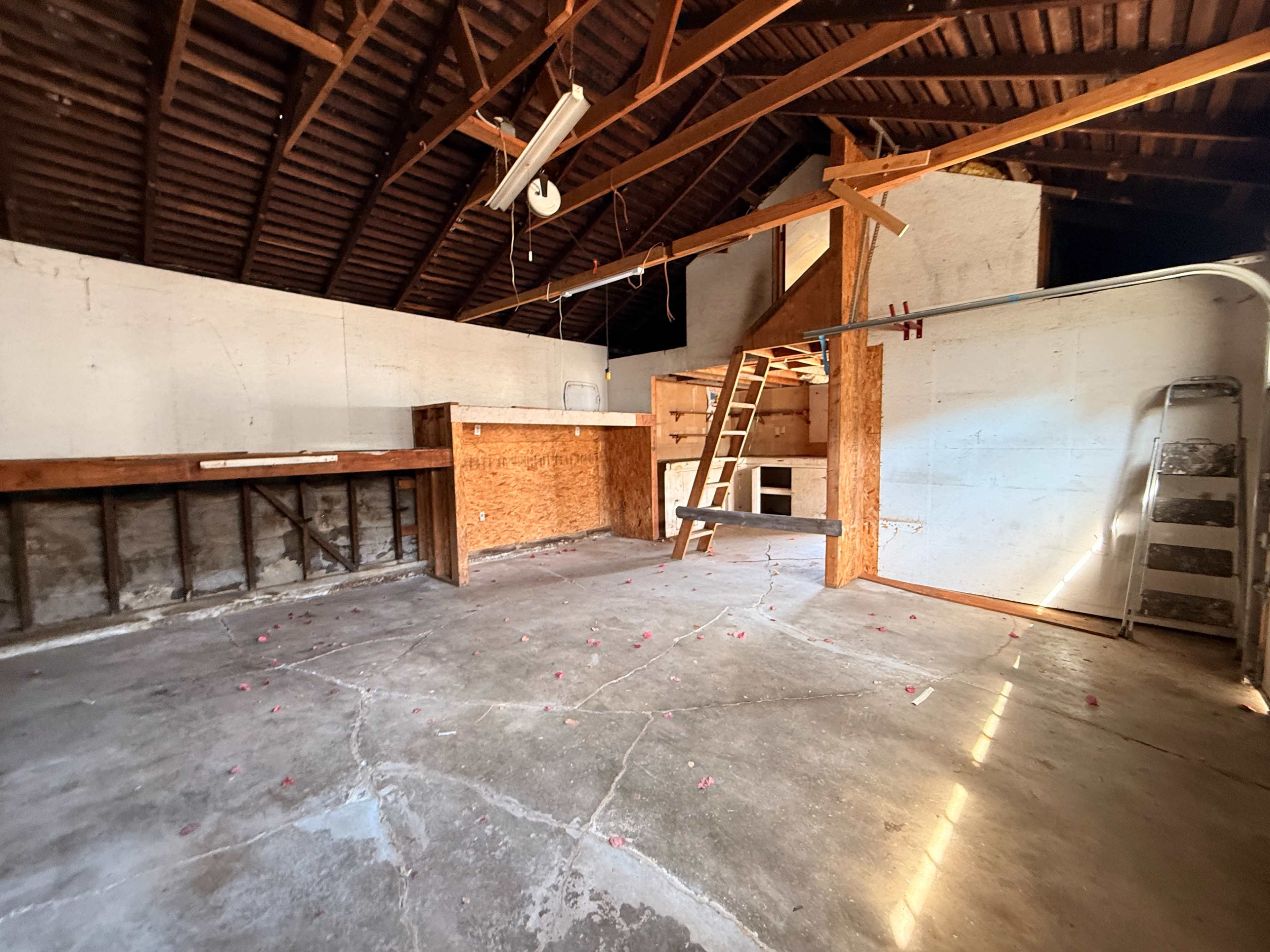 The image shows an unfinished garage or workshop space with exposed beams, concrete flooring, and a wooden ladder leaning against a wall.