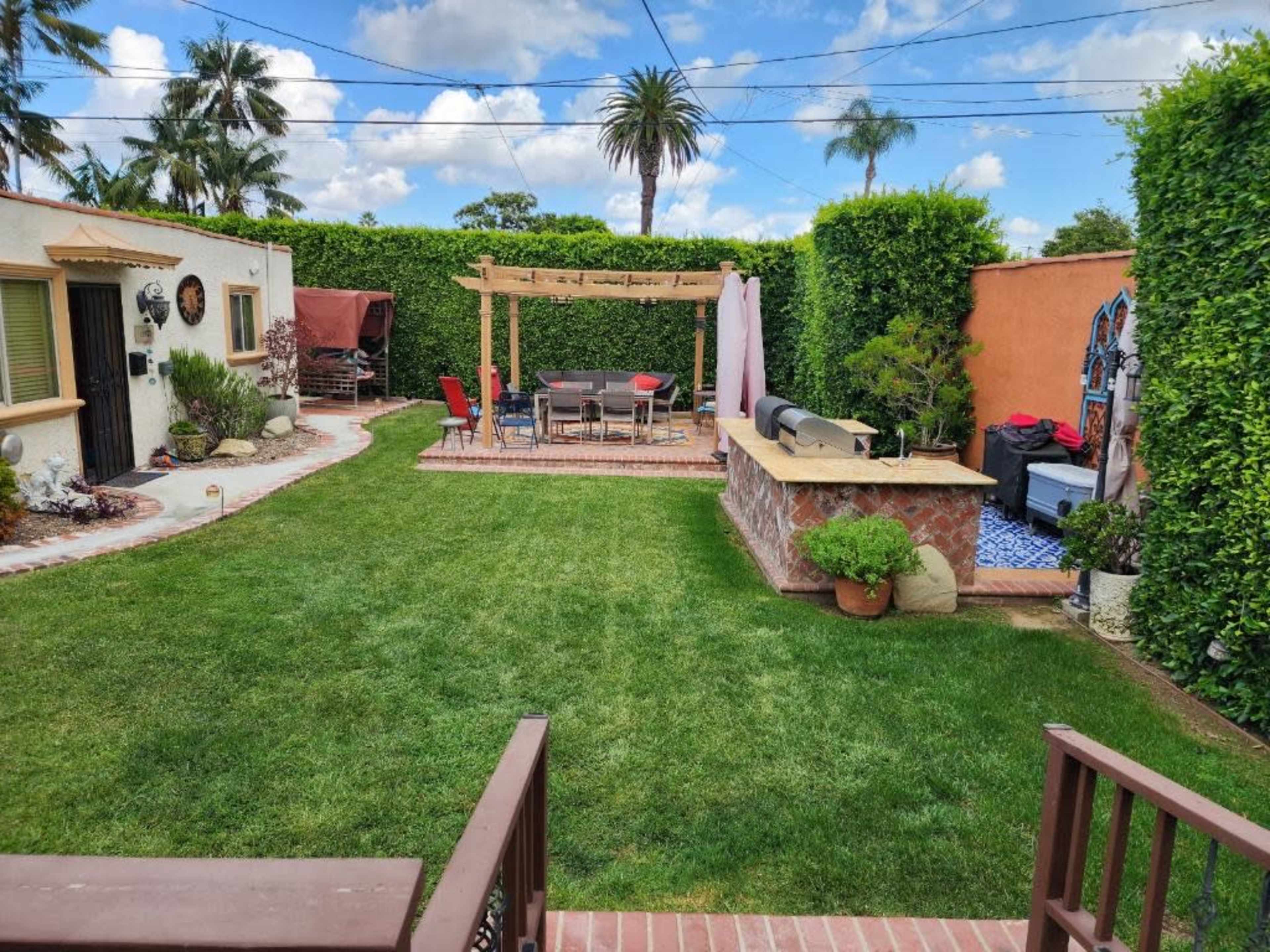 The image shows a landscaped backyard with green grass, a patio area featuring a pergola and dining set, surrounded by tall hedges and palm trees.