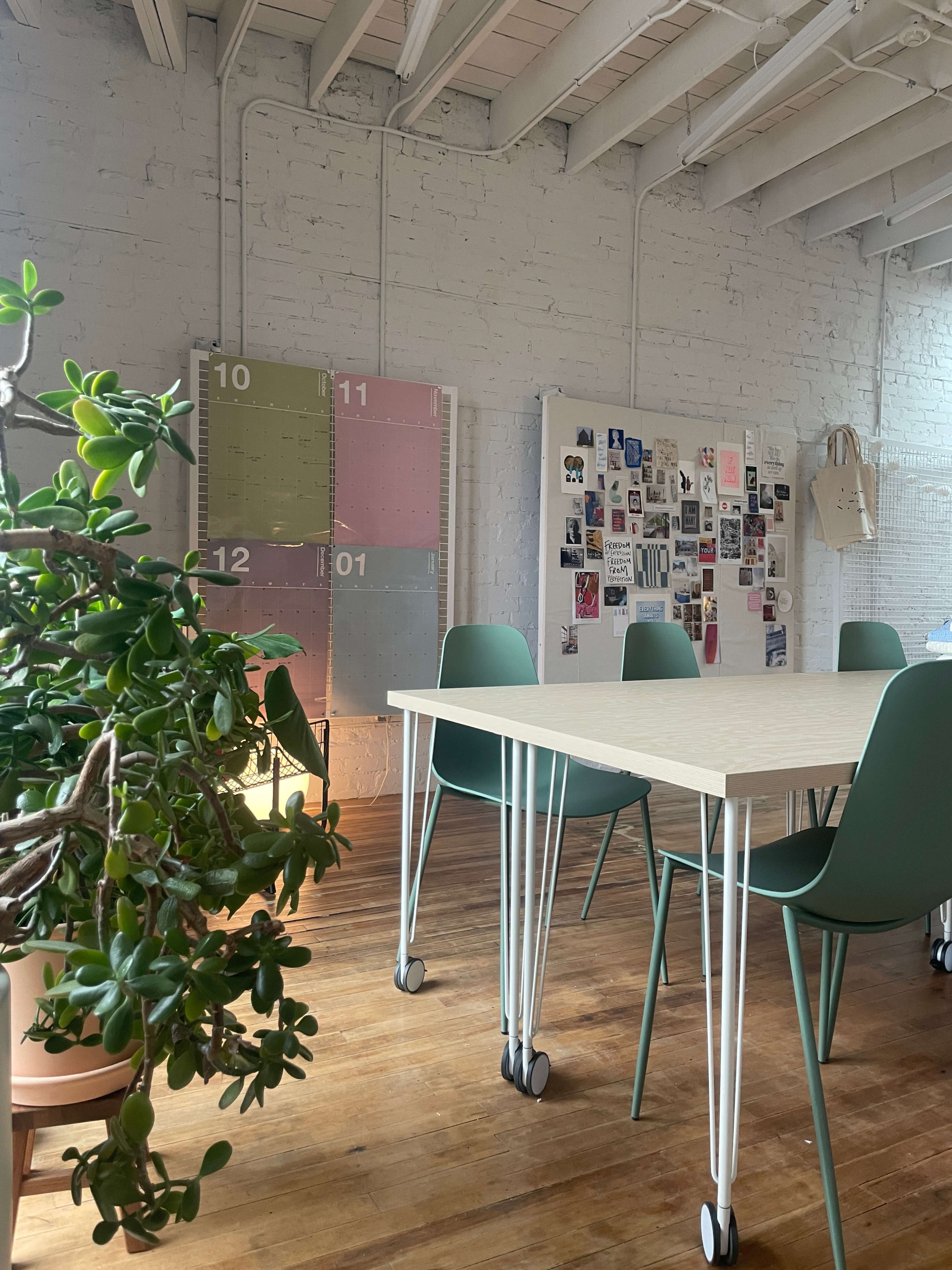 A bright, minimalist workspace features a wooden table with green chairs, a plant nearby, and a wall adorned with colorful calendar pages and a collage of images.