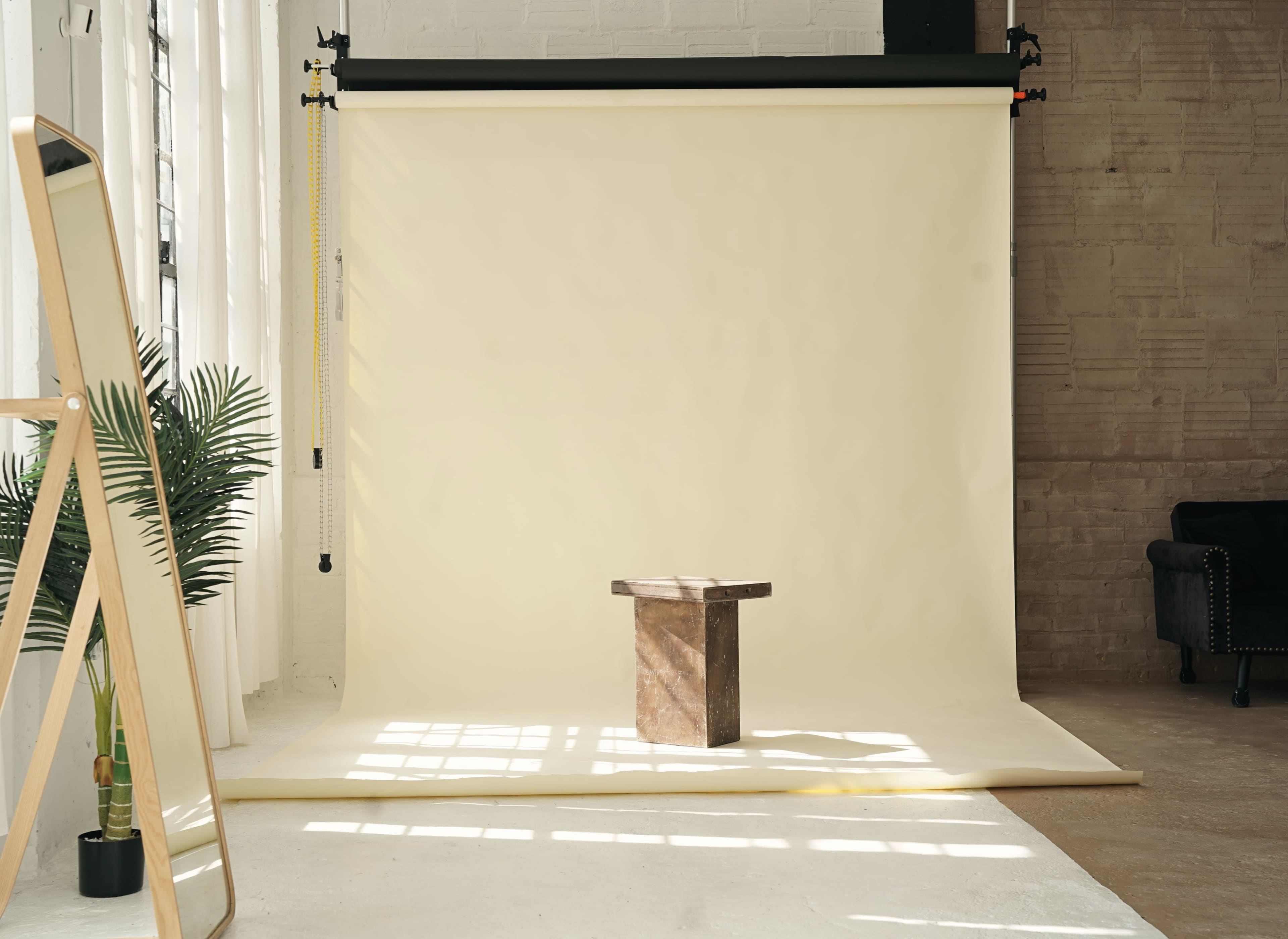 The image shows a minimalist photography studio with a plain beige backdrop and a small wooden table at its center, flanked by a large mirror and a potted plant.