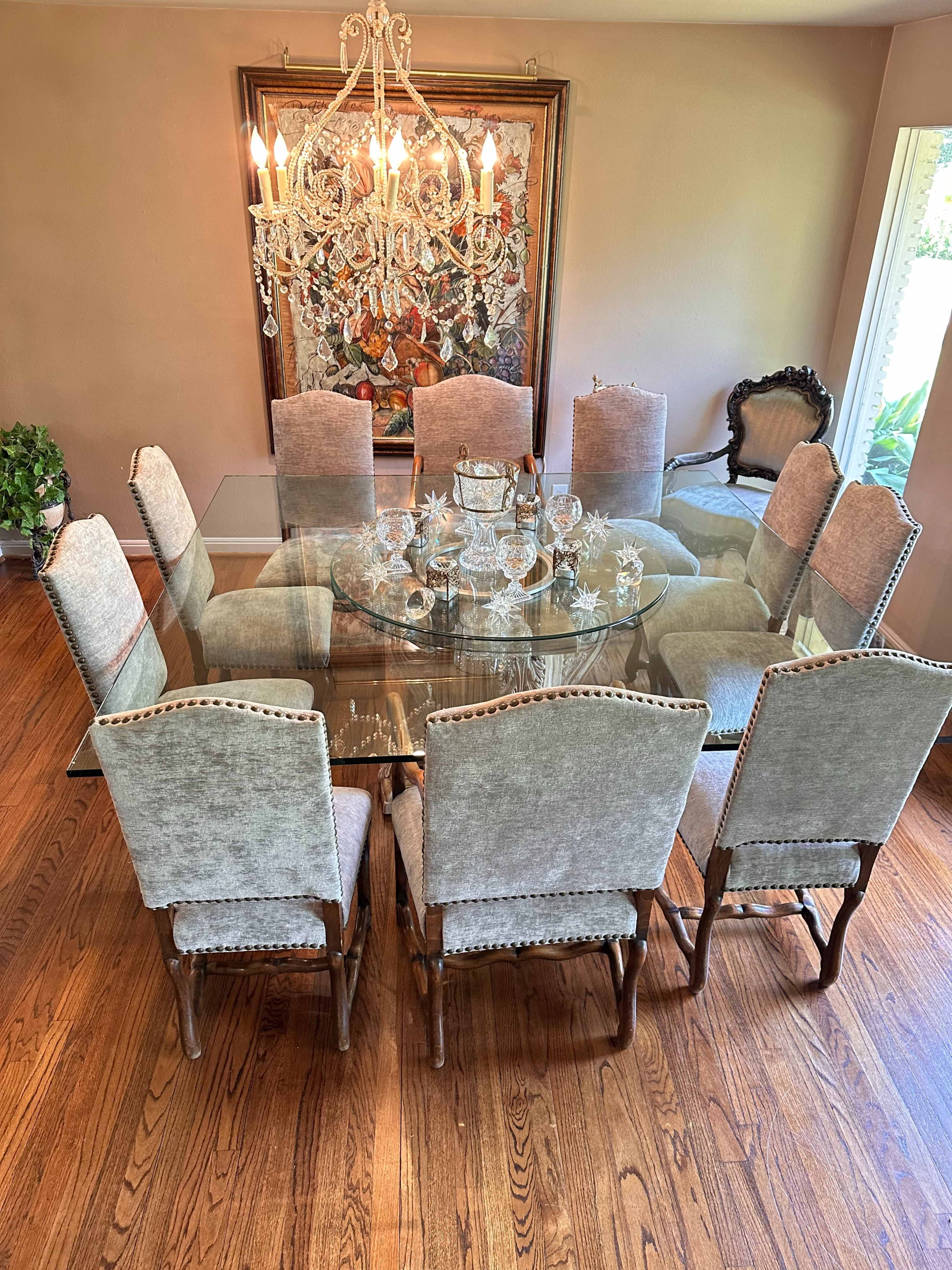 A large, glass dining table surrounded by twelve upholstered chairs is illuminated by a crystal chandelier in a well-lit room.