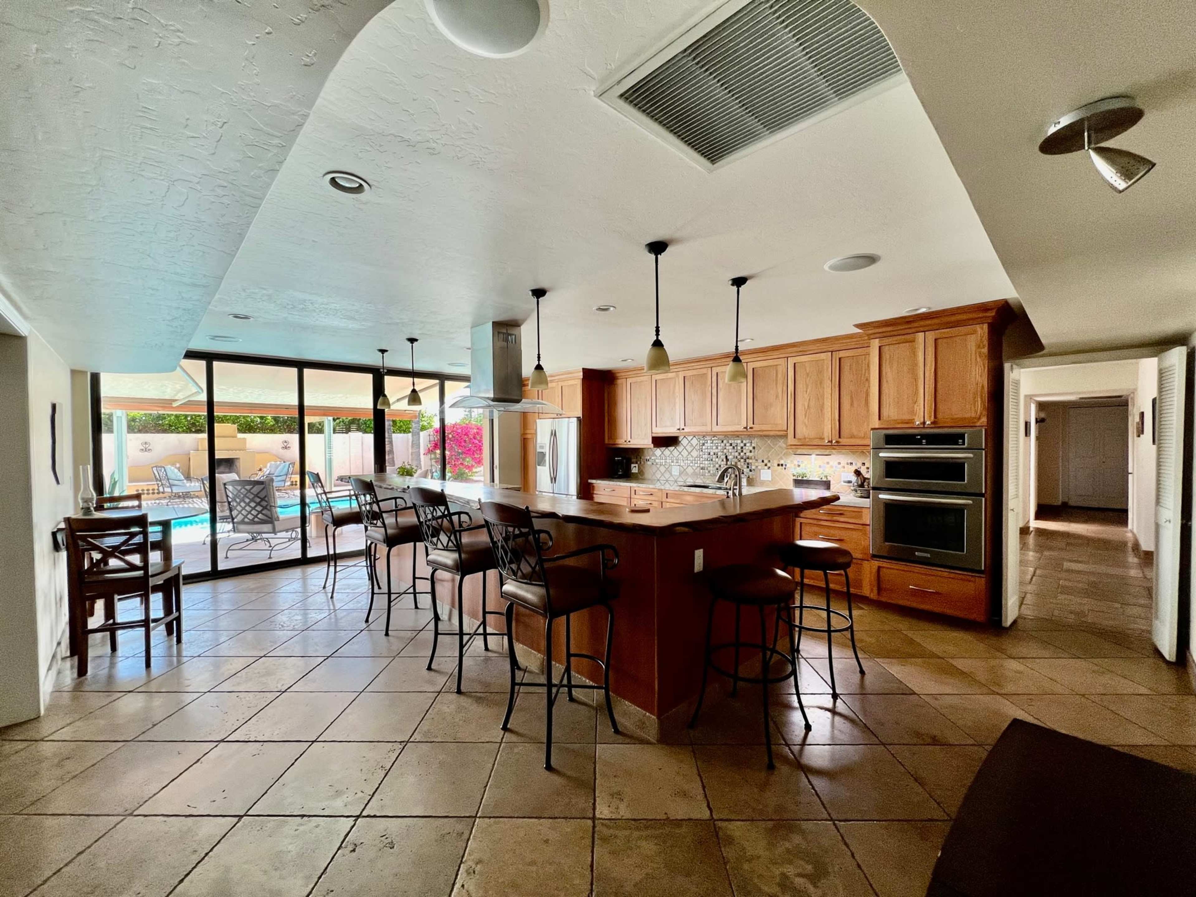 A modern kitchen with wooden cabinetry, an island with bar seating, and large windows overlooking a pool area.