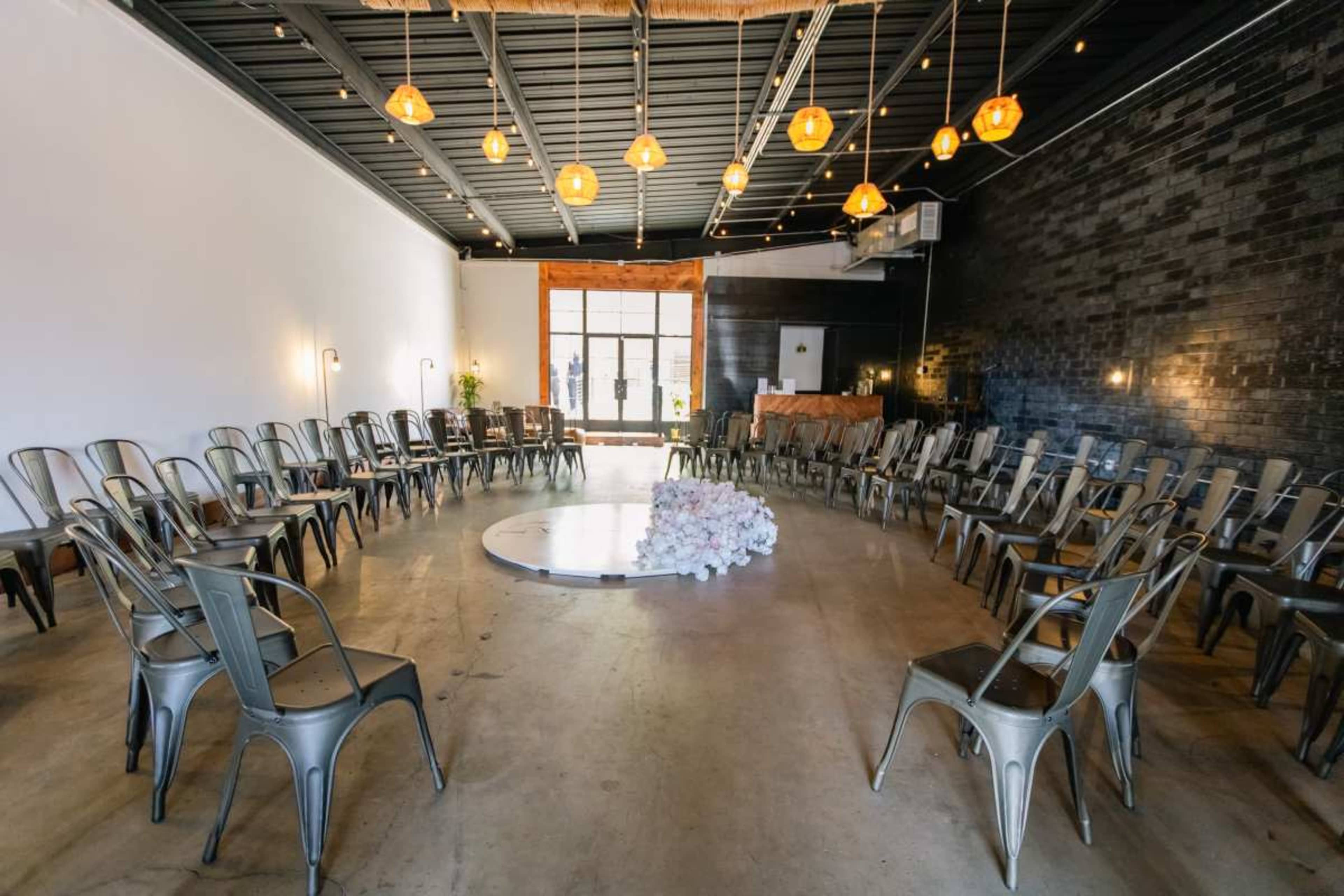 A circular arrangement of metal chairs encompasses a white circular platform in a well-lit event space with a dark brick wall.