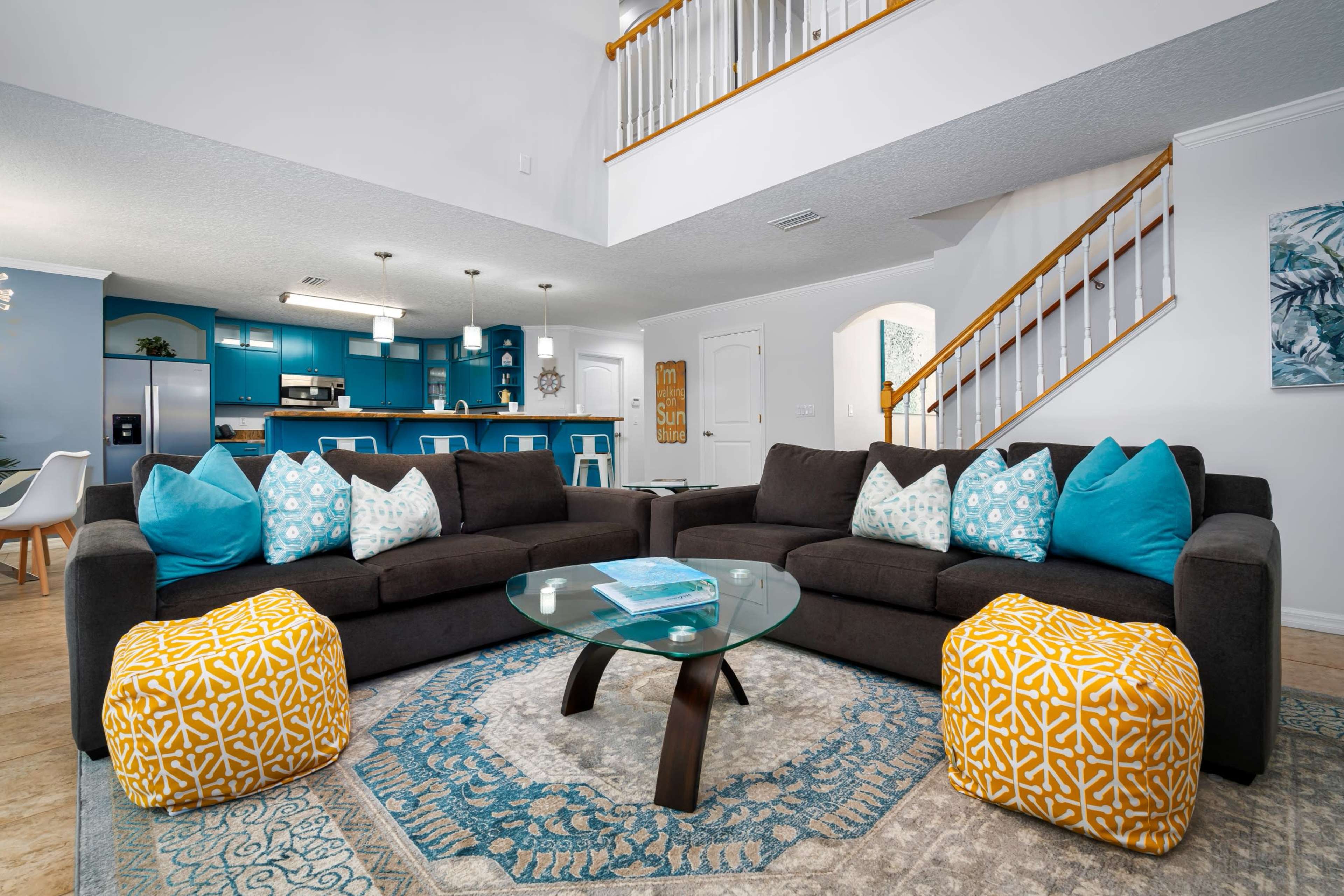 A spacious living area features a dark sectional sofa with colorful cushions, a glass coffee table, and decorative poufs, adjacent to a modern kitchen with blue cabinetry.