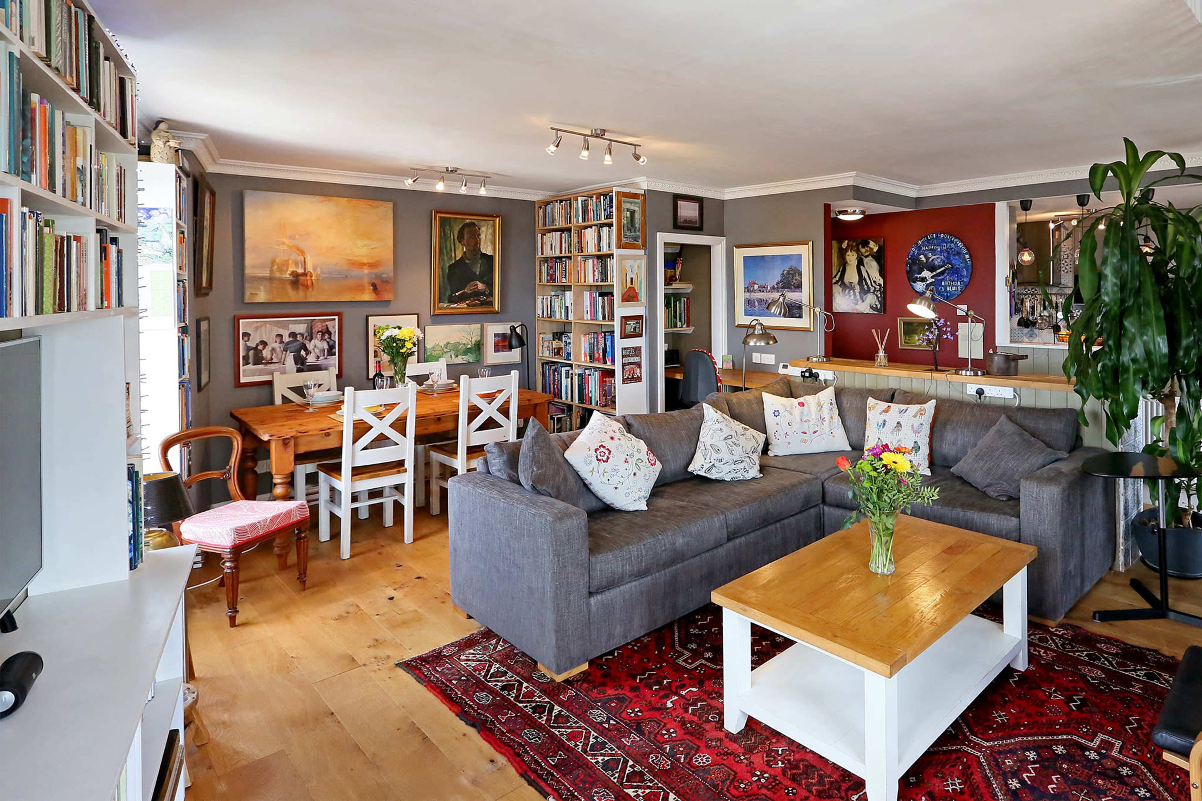 The scene shows a cozy living room with a gray sectional sofa, a wooden dining table, bookshelves filled with books, and various artworks on the walls.