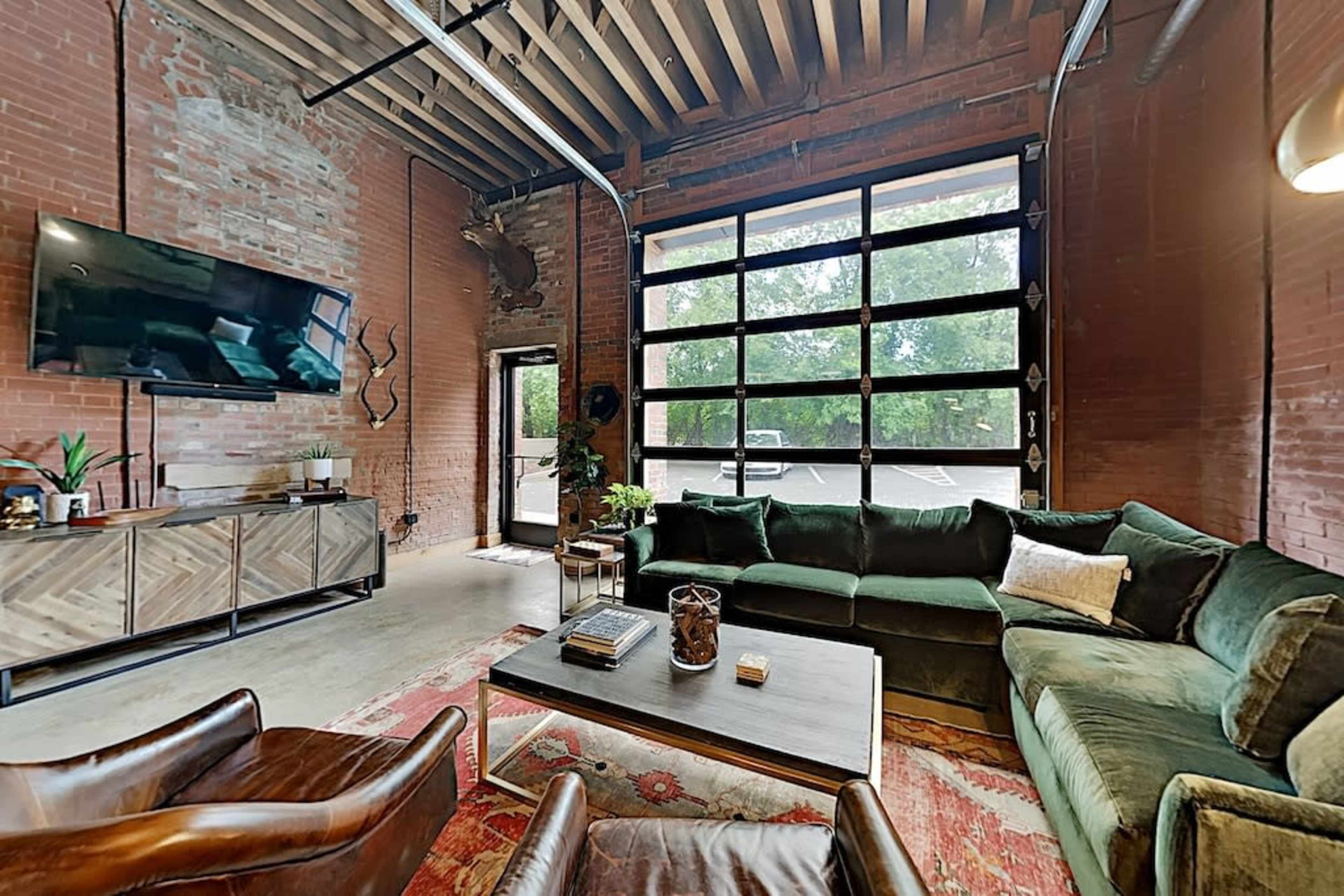 A spacious living area with exposed brick walls, large windows, and a green sectional sofa facing a coffee table.
