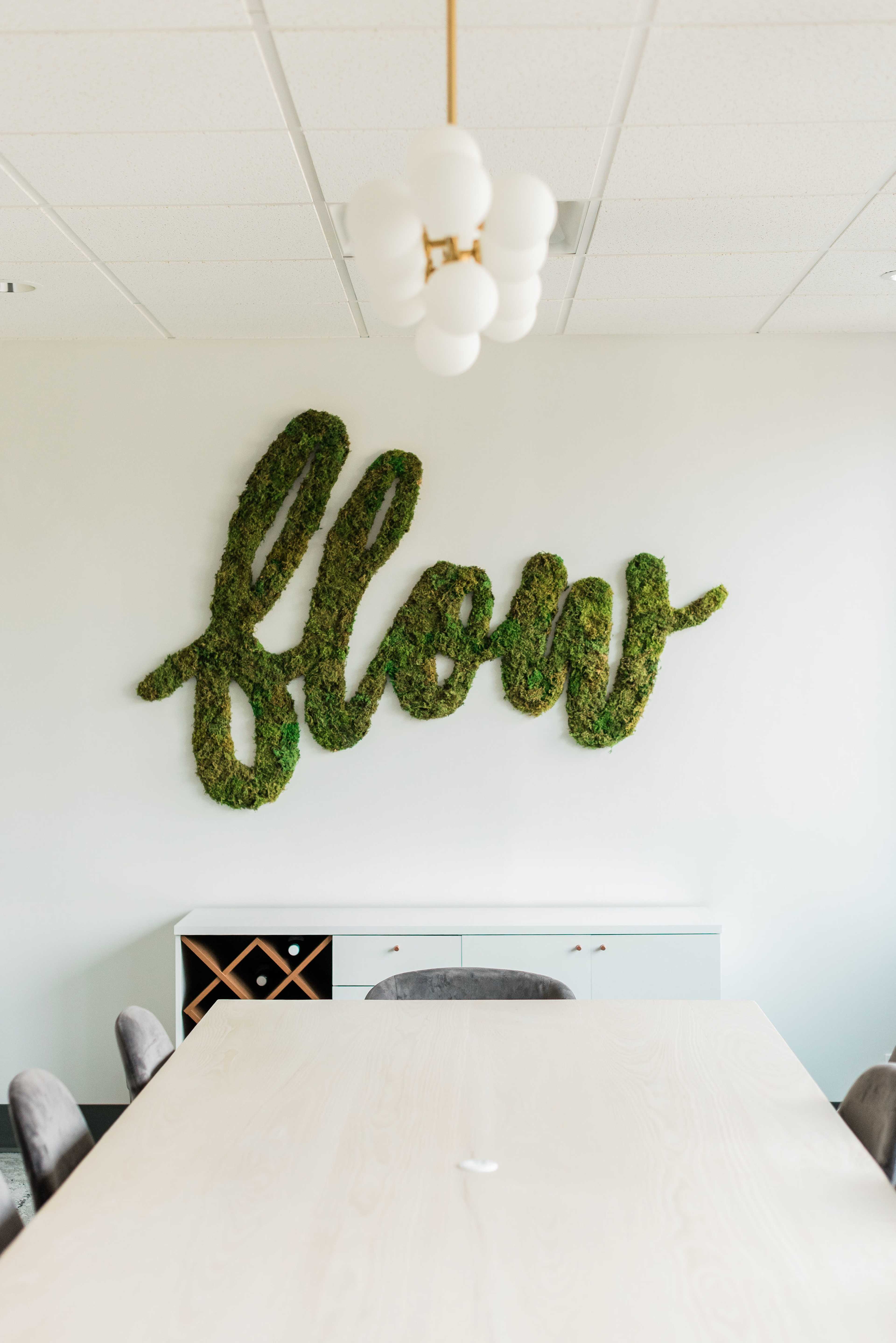 A moss-covered word "flow" is displayed on a white wall above a light-colored wooden table in a modern office setting.