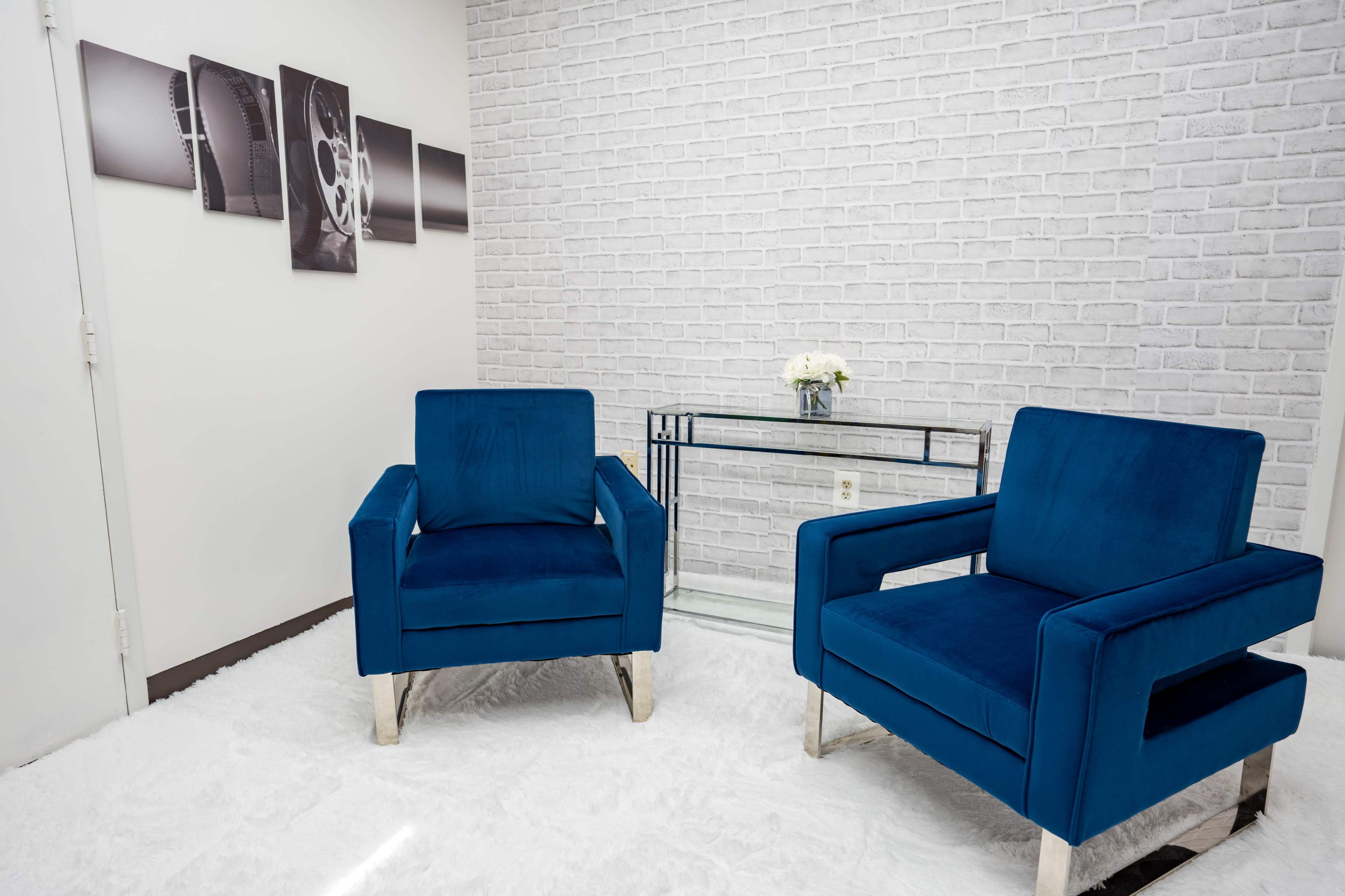 A modern sitting area with two blue chairs facing a glass table, situated against a white brick wall.
