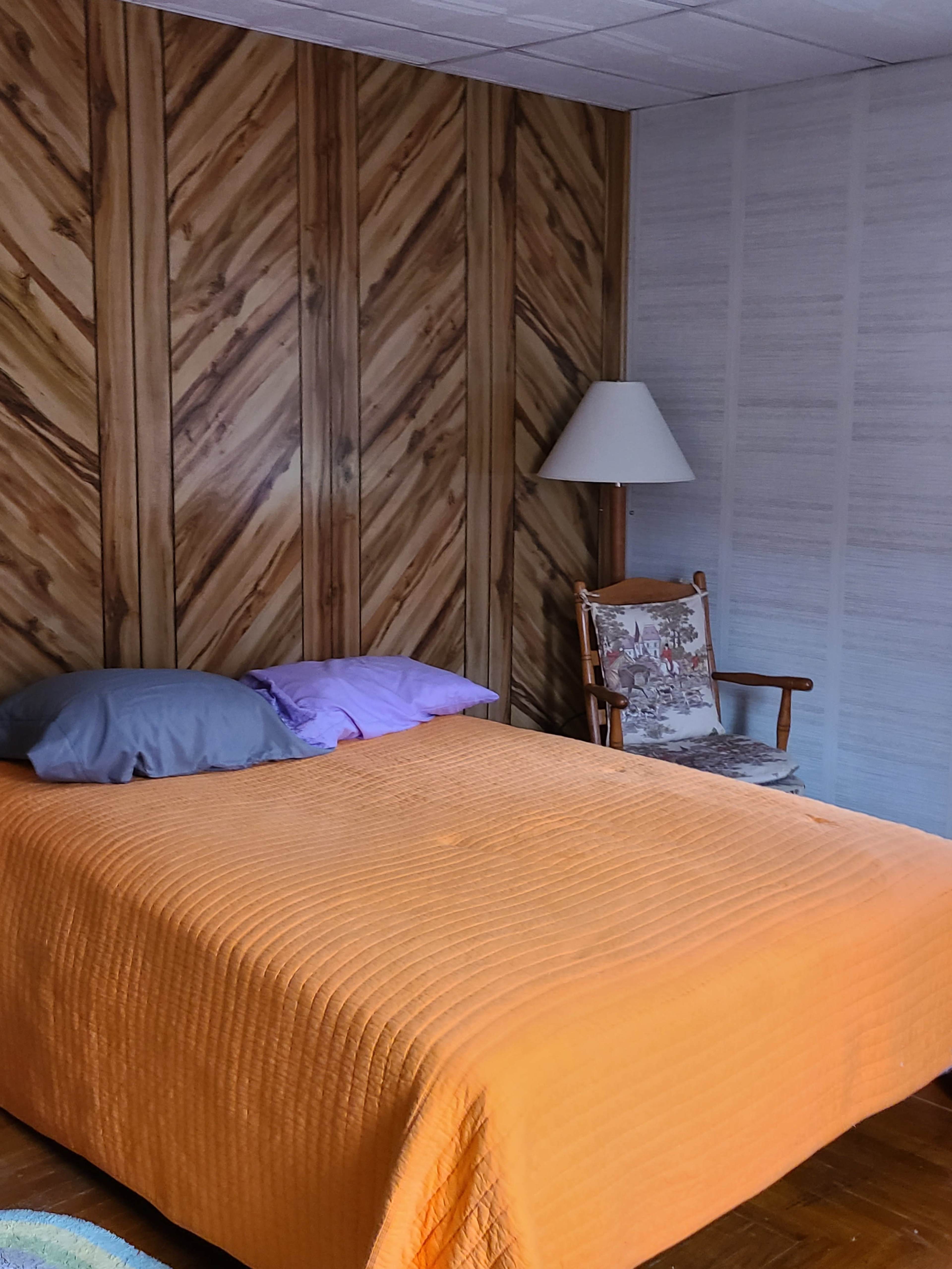 A simple bedroom with a bed covered in an orange quilt, two pillows, a wooden chair, and a lamp against a patterned wall.