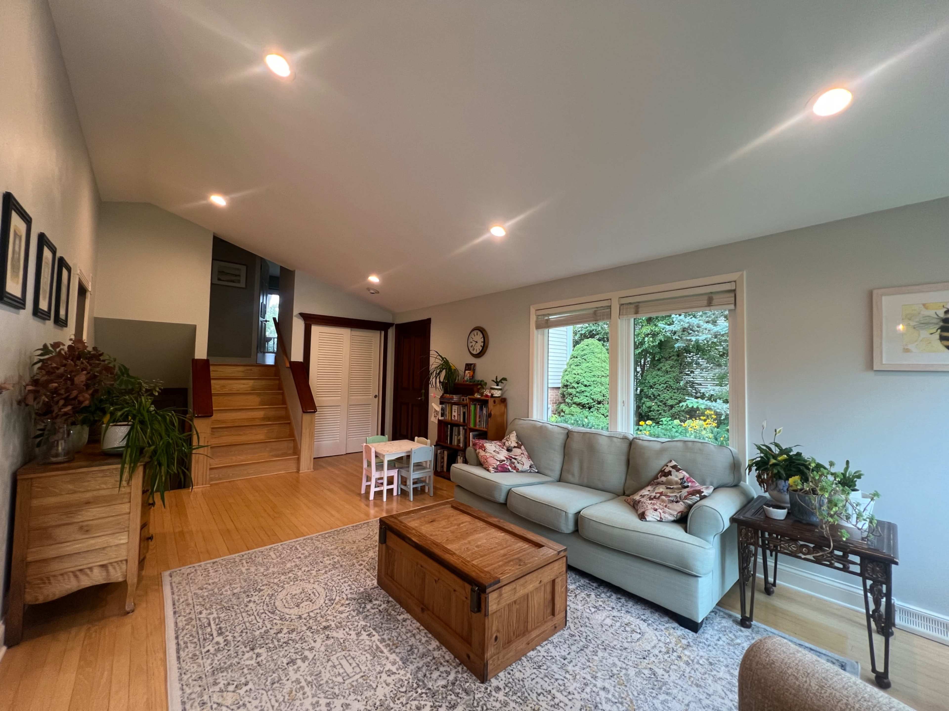 The image shows a bright living room with a light blue sofa, a wooden coffee table, and a staircase leading to another level, adorned with plants and decorative elements.