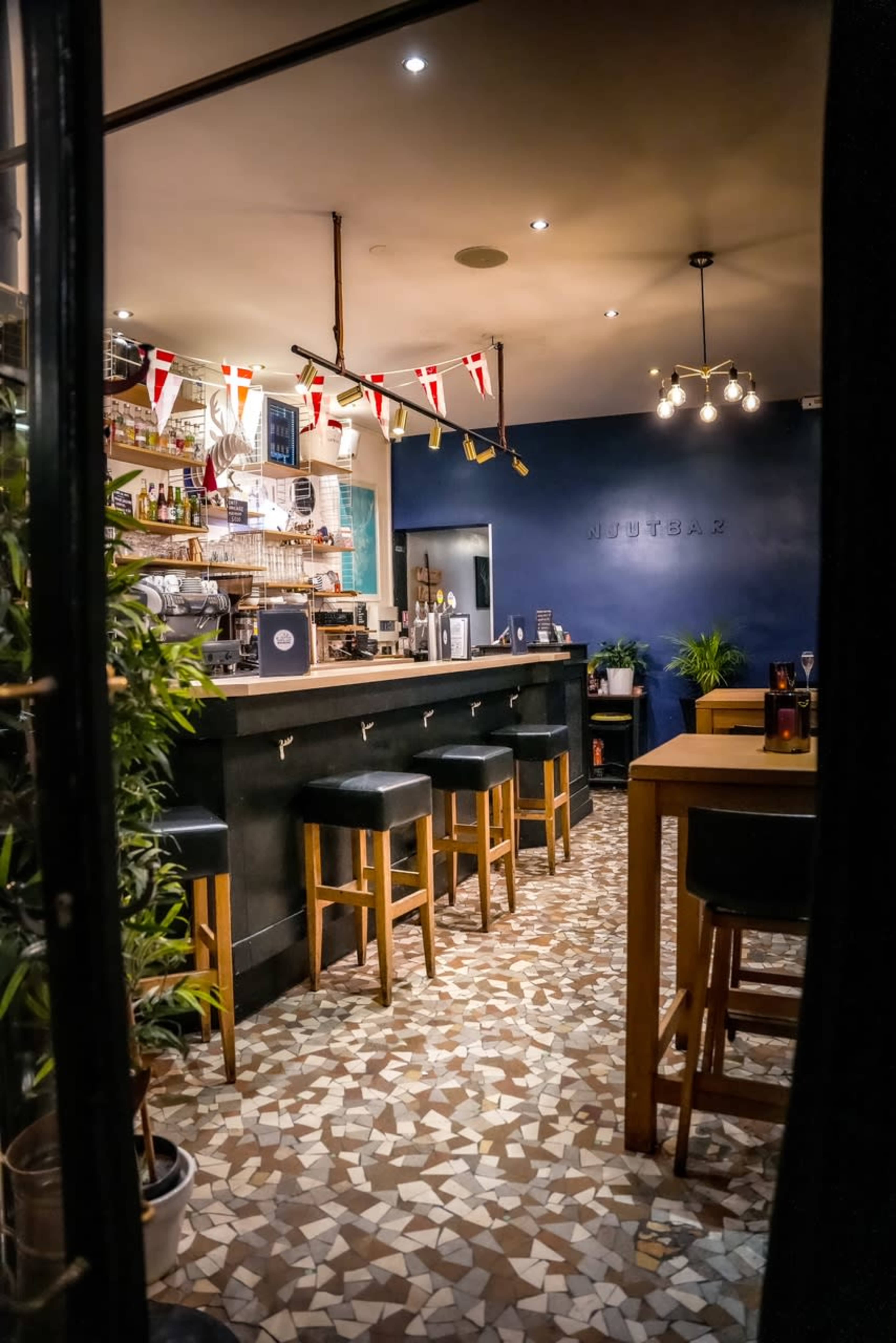 A modern bar area with a decorated counter, high stools, and flag bunting hanging above, set against a backdrop of tiled flooring and blue walls.