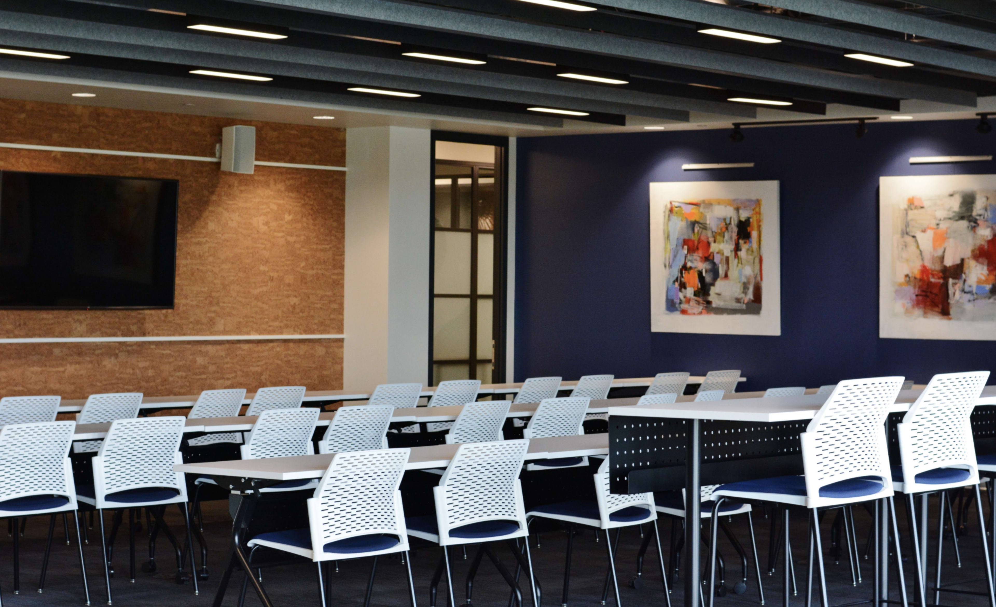 A modern meeting room features rows of white chairs and a long table, with colorful artwork on the walls and a large screen mounted.