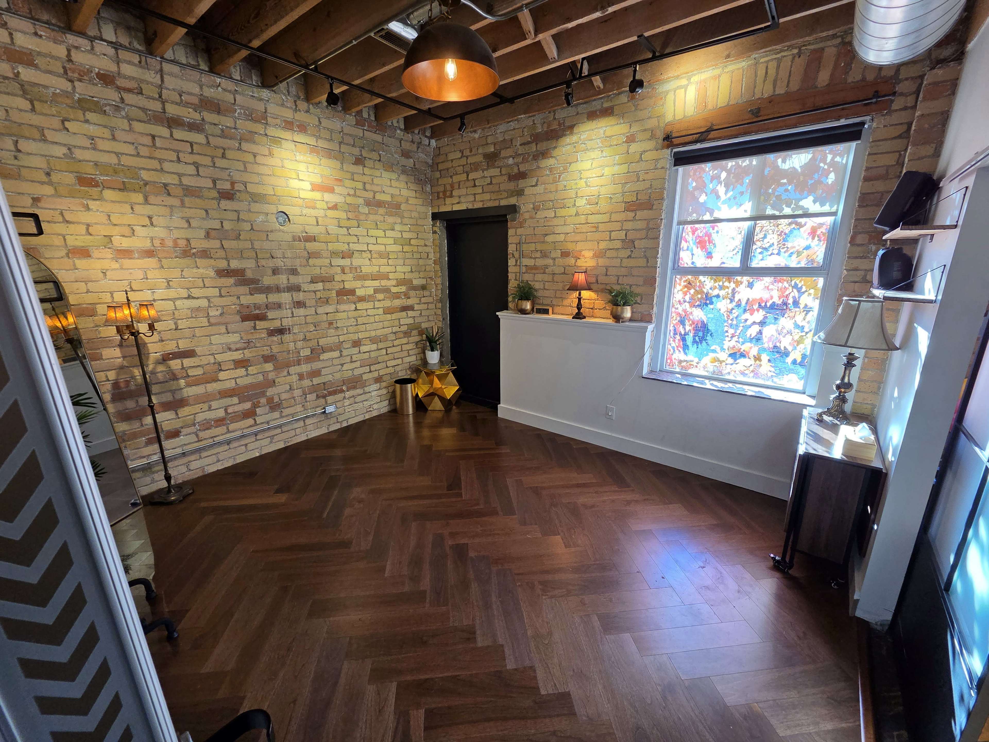 The image shows a spacious room with exposed brick walls, hardwood herringbone flooring, and a large window featuring colorful autumn foliage outside.