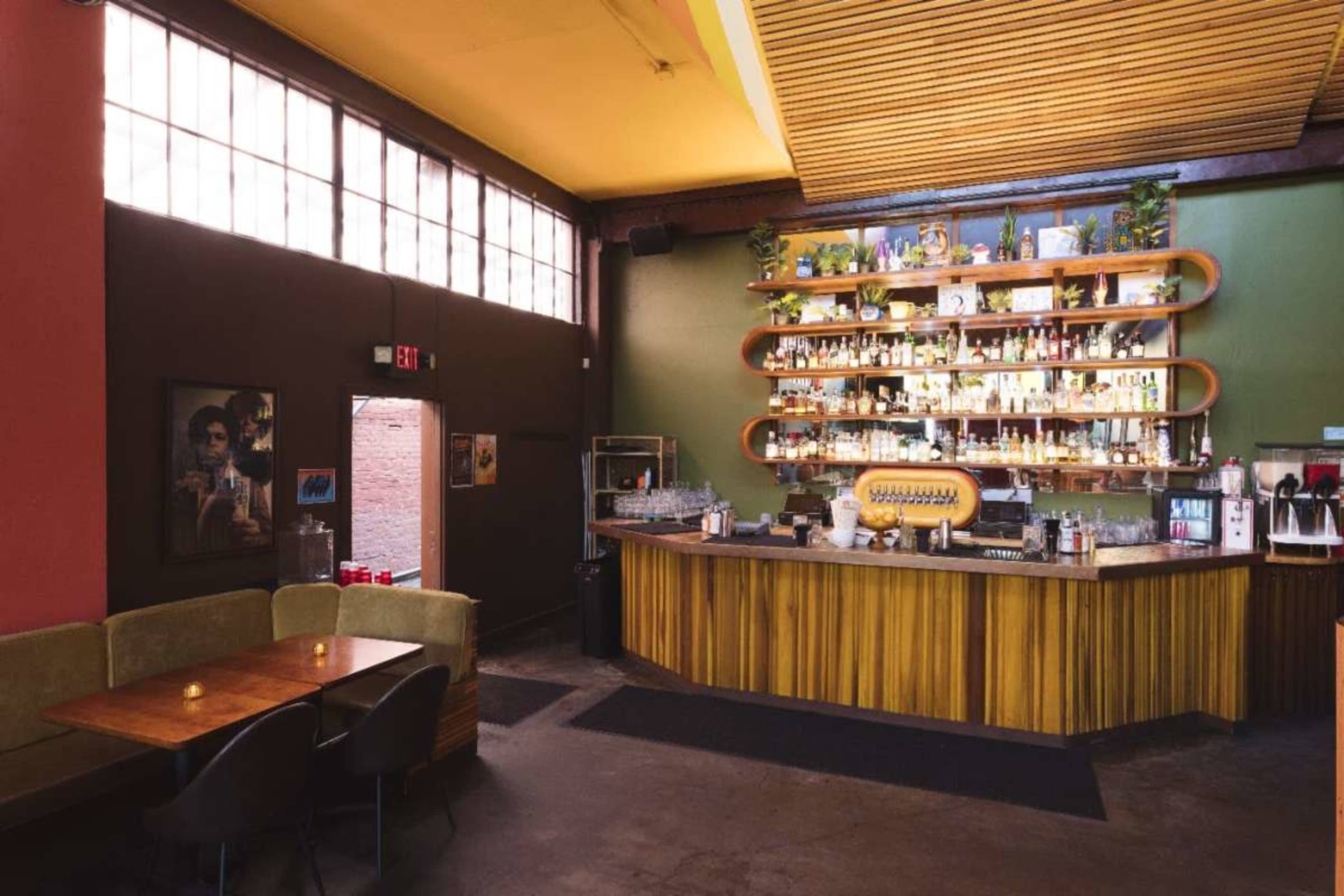 The image shows a bar interior featuring a well-stocked shelf of liquor bottles, a wooden bar counter, and seating areas with muted lighting.