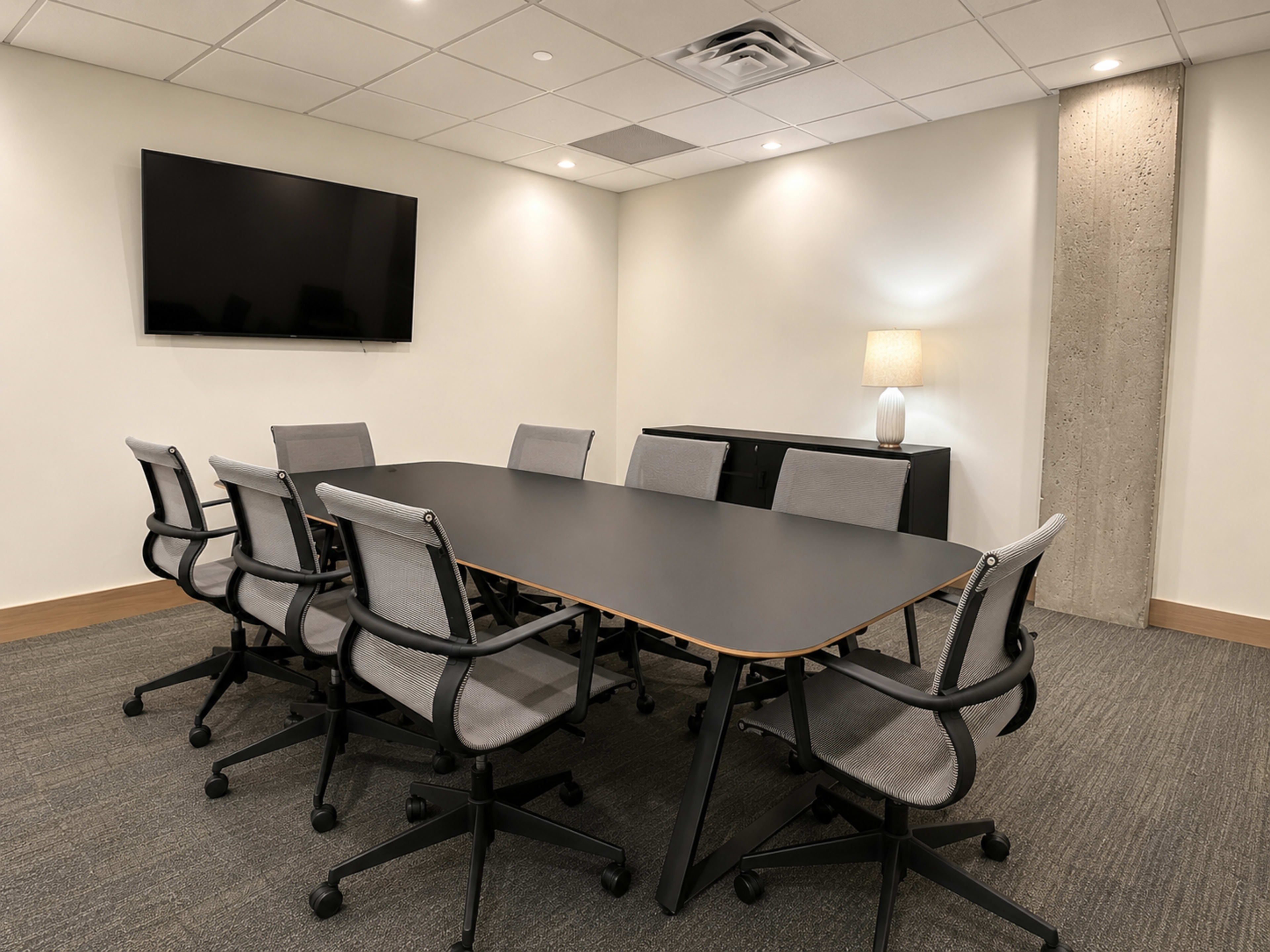 A modern conference room featuring a long black table surrounded by ergonomic chairs, with a flat-screen TV mounted on the wall and a side cabinet.