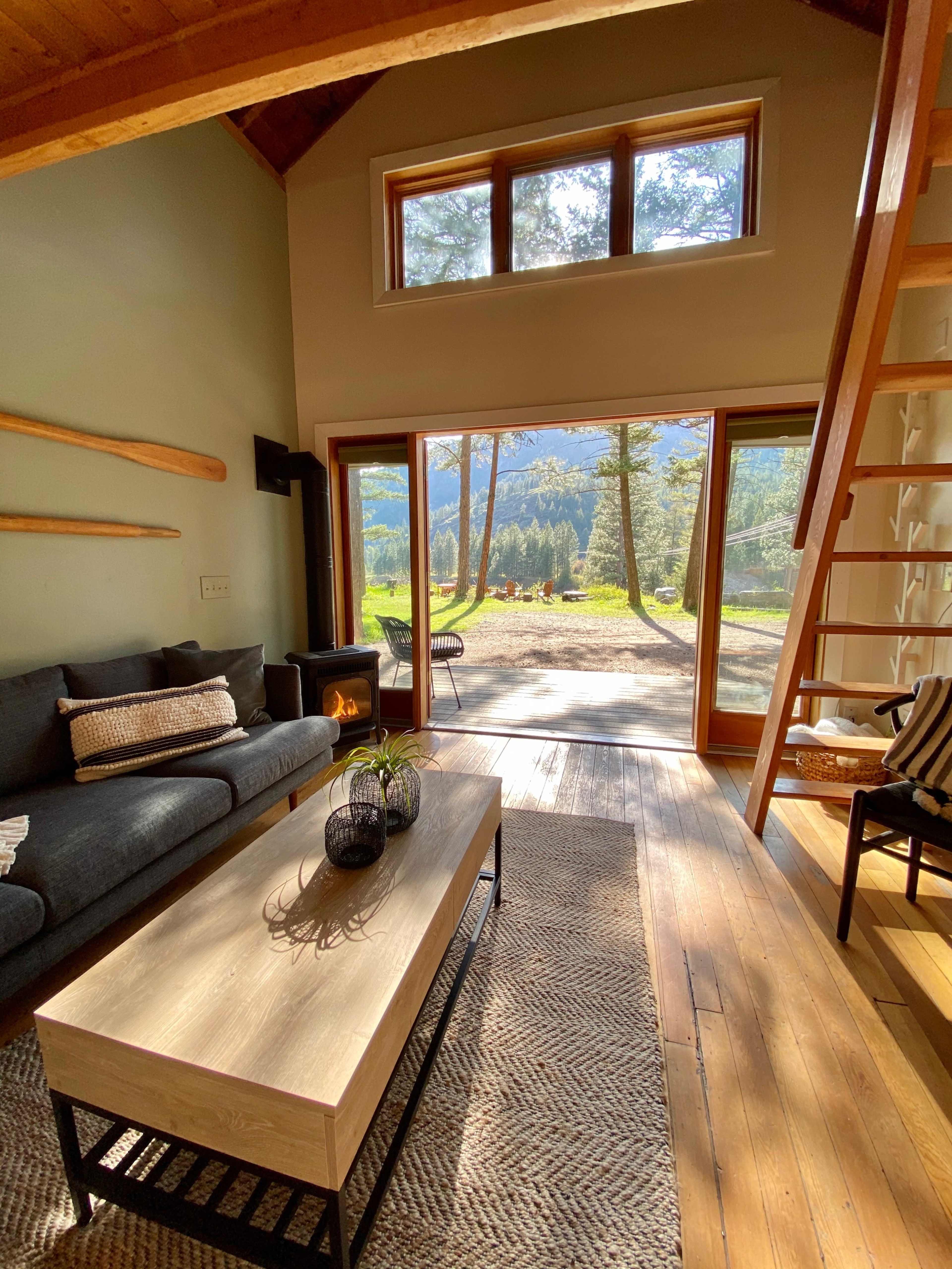 A cozy living room with a wooden ladder leading to a loft, a coffee table in the center, and large windows showcasing a view of trees and mountains outside.
