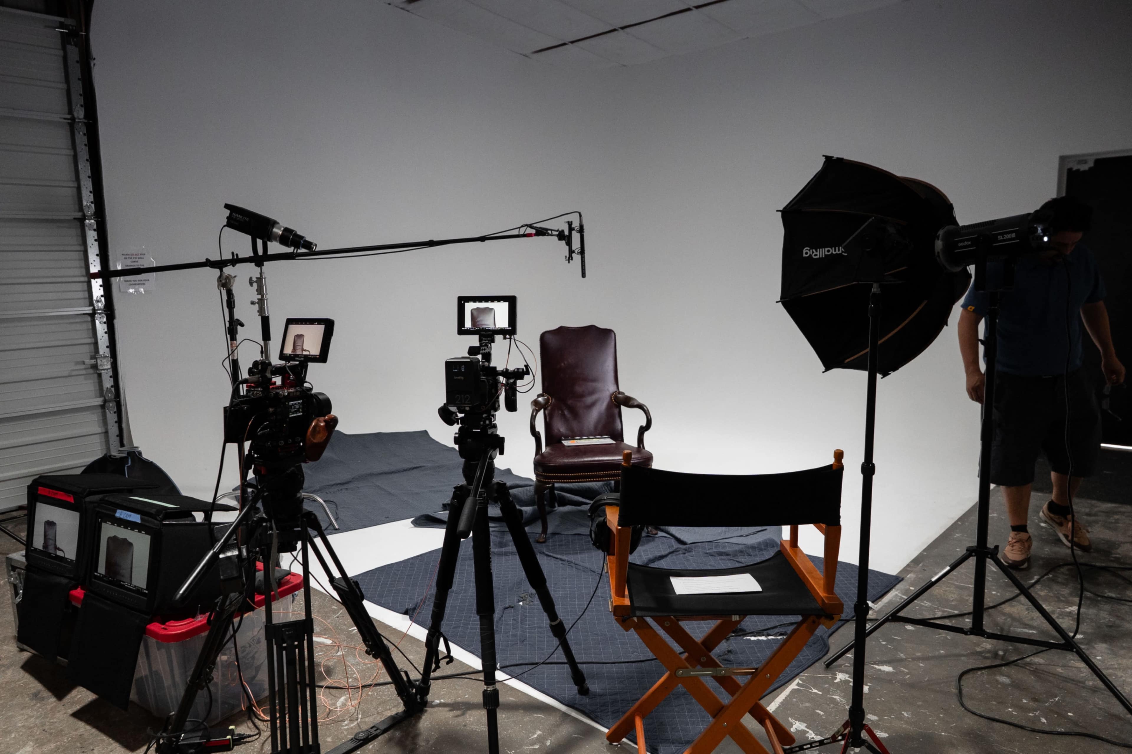 A photography studio is set up with two cameras facing an empty chair, surrounded by lighting equipment and a backdrop.