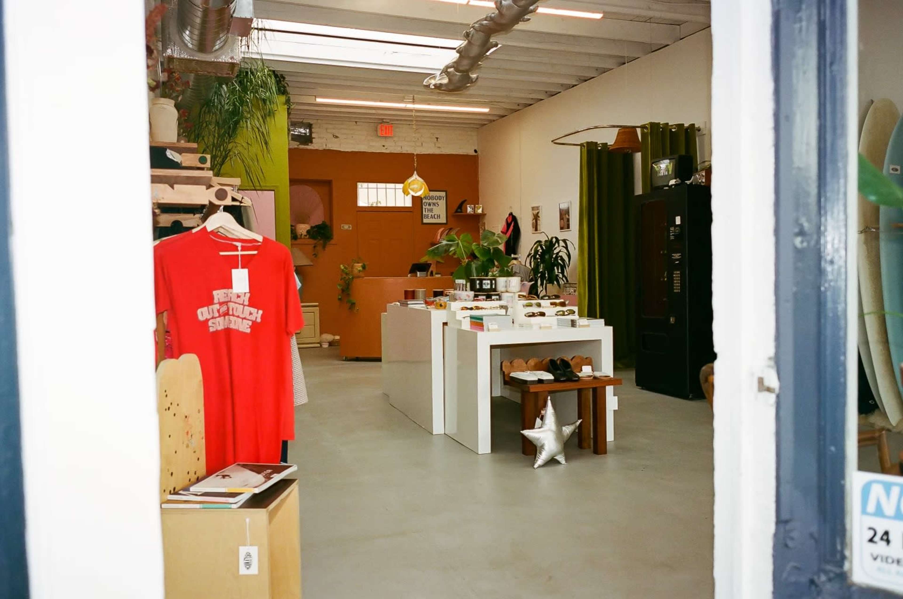 The interior of a retail space featuring a red t-shirt on display, a white product table, various plants, and minimalist decor.