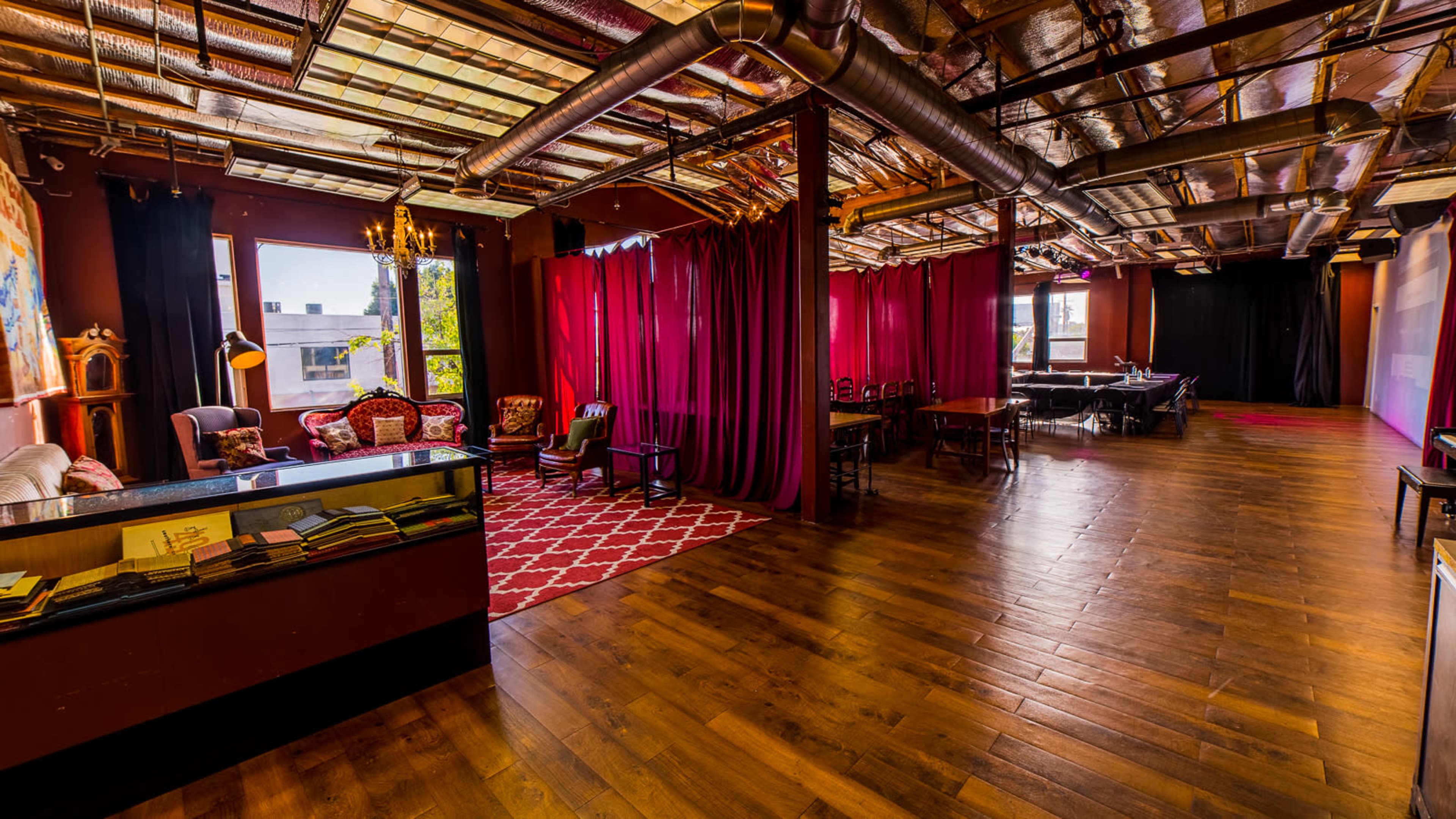 A spacious interior with wooden flooring, featuring a seating area with red curtains, vintage furniture, and large windows allowing natural light to enter.
