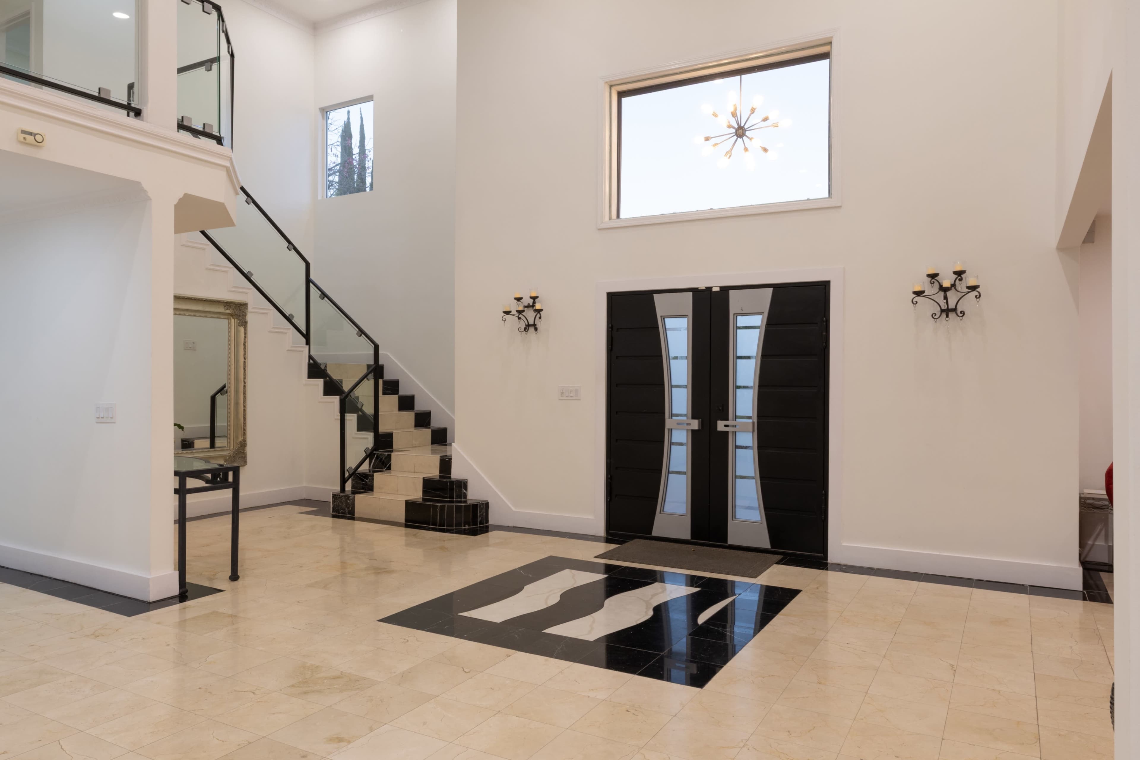 The image shows a spacious foyer with a modern staircase, elegant black double doors, and a polished marble floor featuring a monochromatic design.