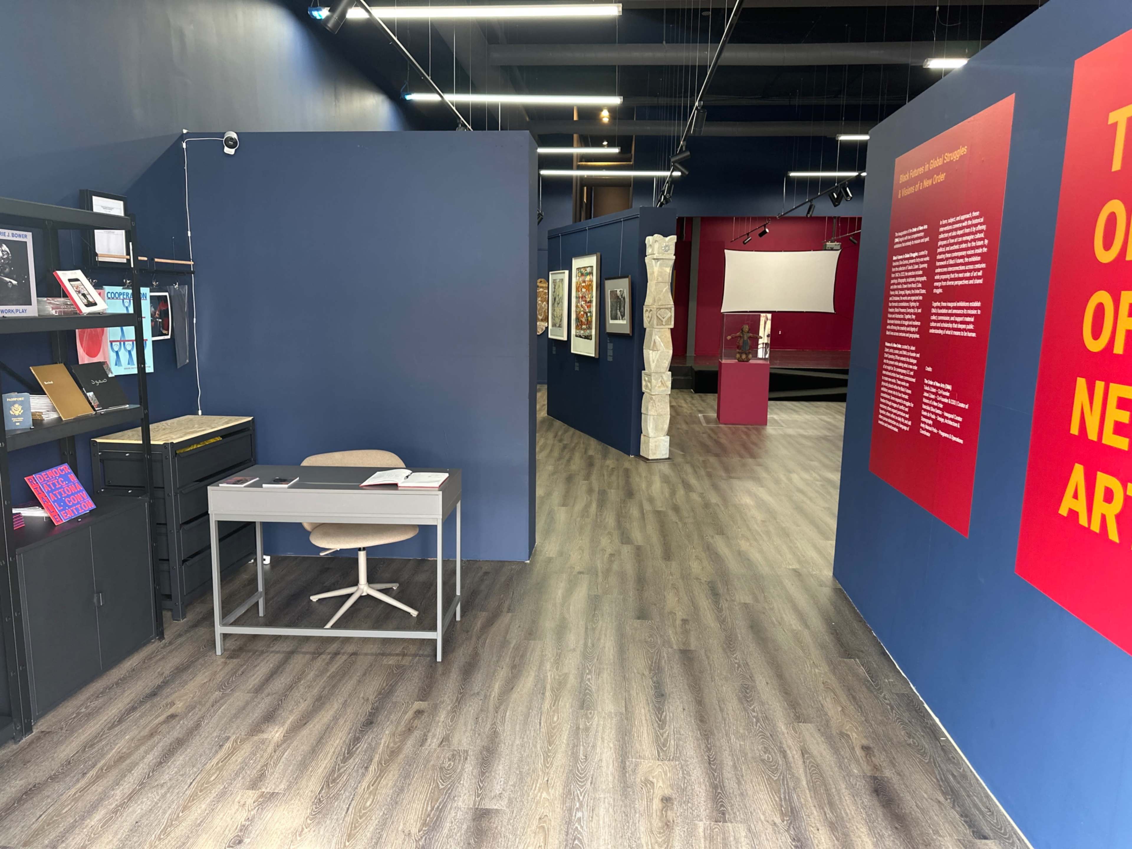 The image shows an art gallery interior with a reception area on the left, featuring a desk and display shelves, and a pathway leading into exhibition spaces with framed artworks on the walls.