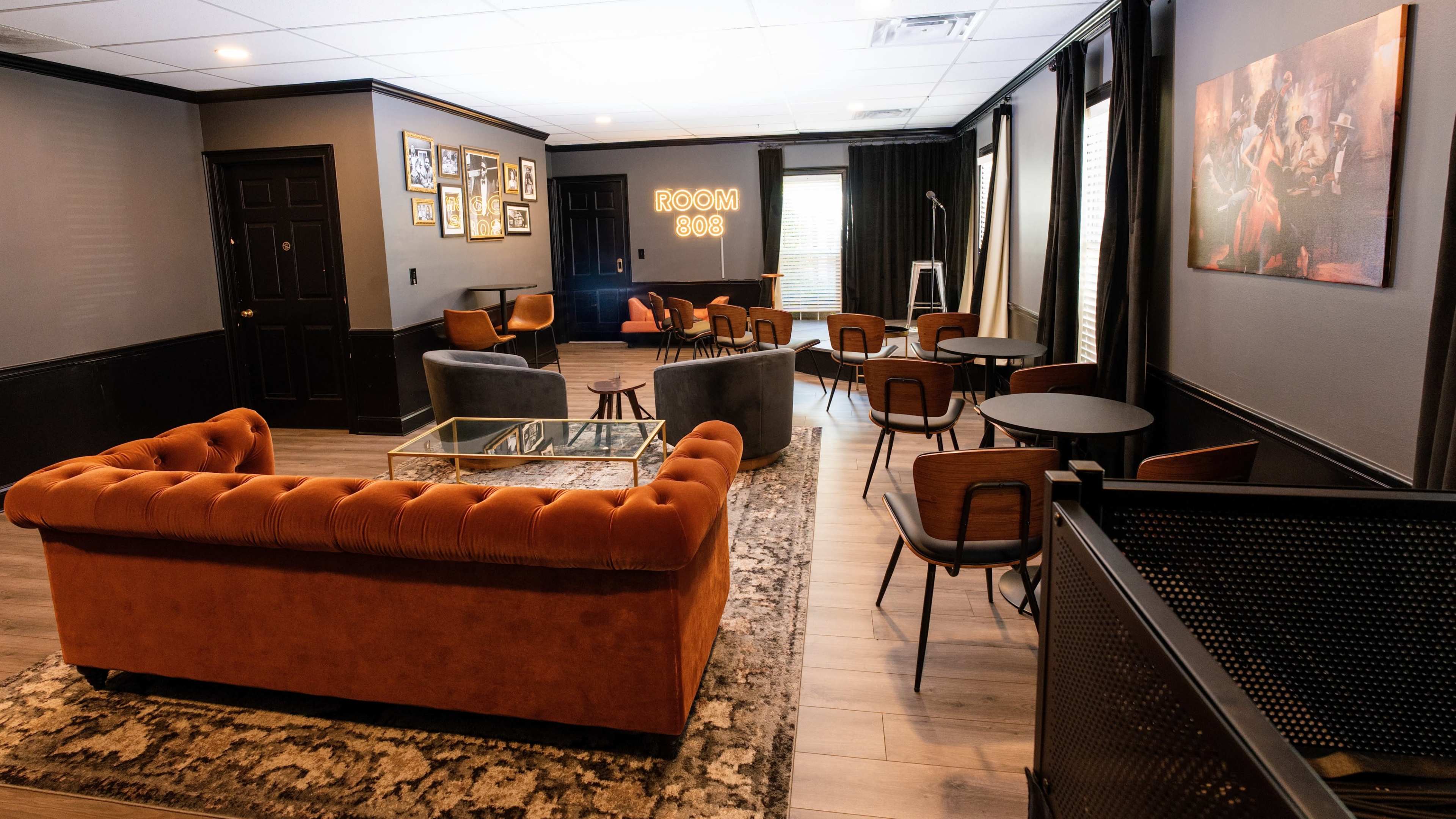 The image shows a modern lounge area featuring a mix of seating arrangements, including an orange tufted sofa, gray armchairs, and wooden tables, with art on the walls.