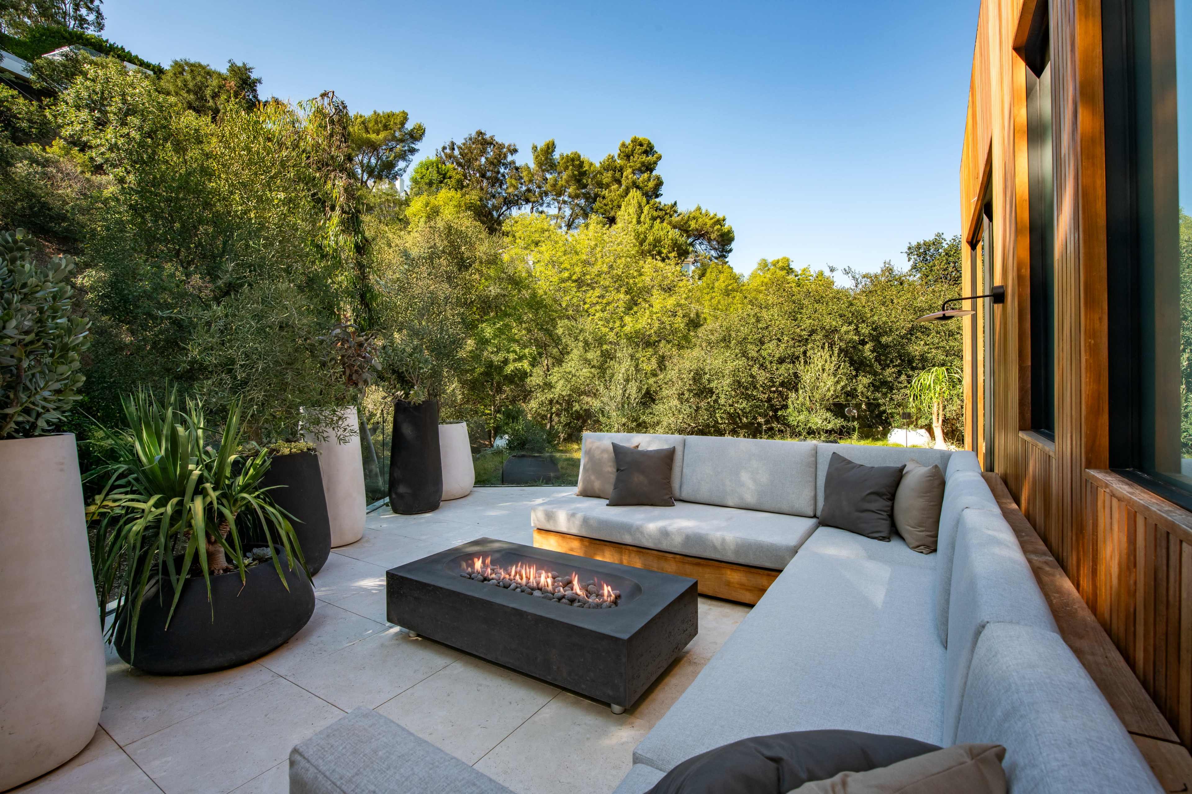 A spacious outdoor lounge area features a gray sectional sofa and a modern fire pit surrounded by lush greenery.