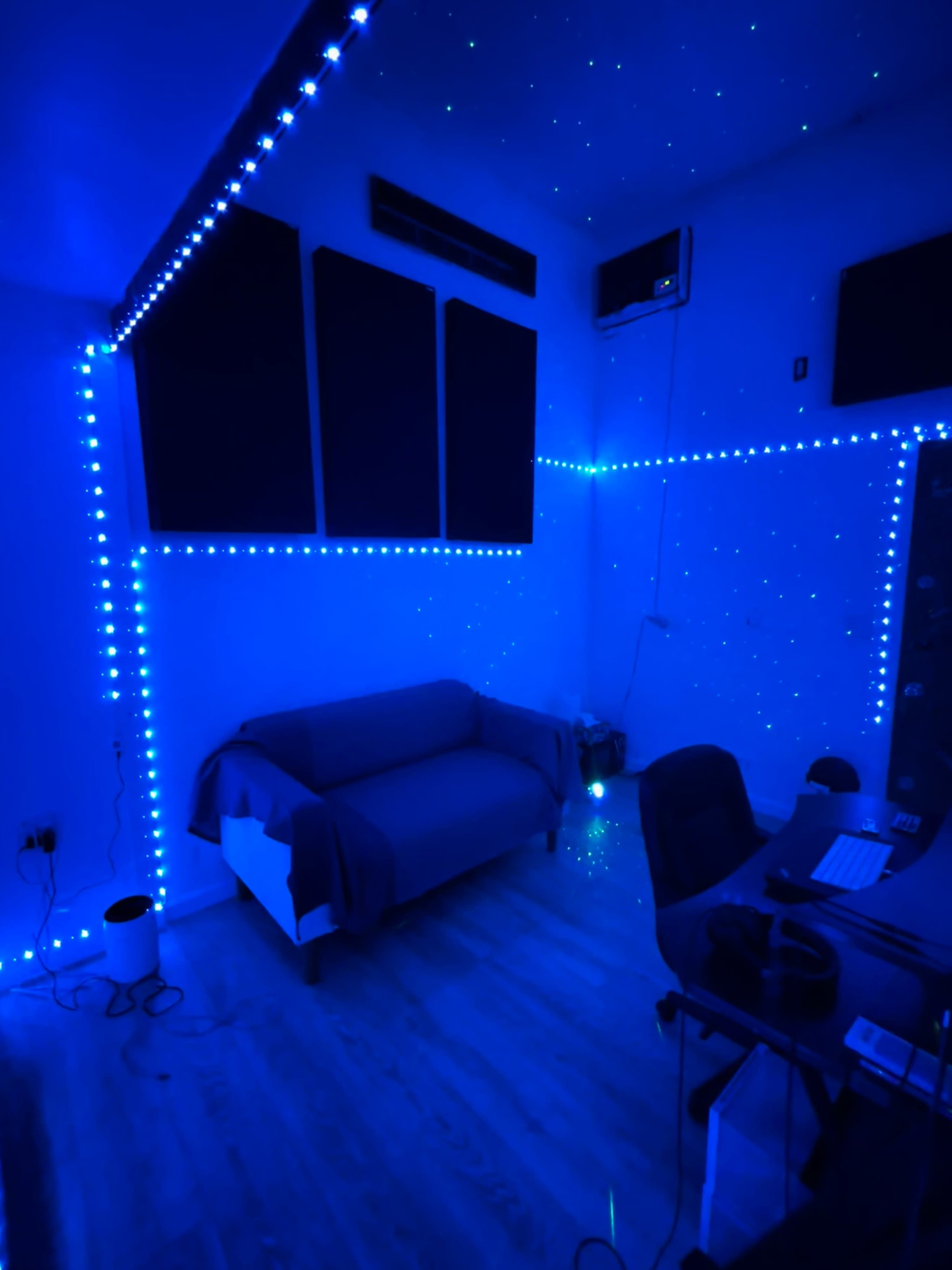 The room is illuminated with blue LED lights, featuring a sofa, a desk with a computer, and acoustic panels on the walls.