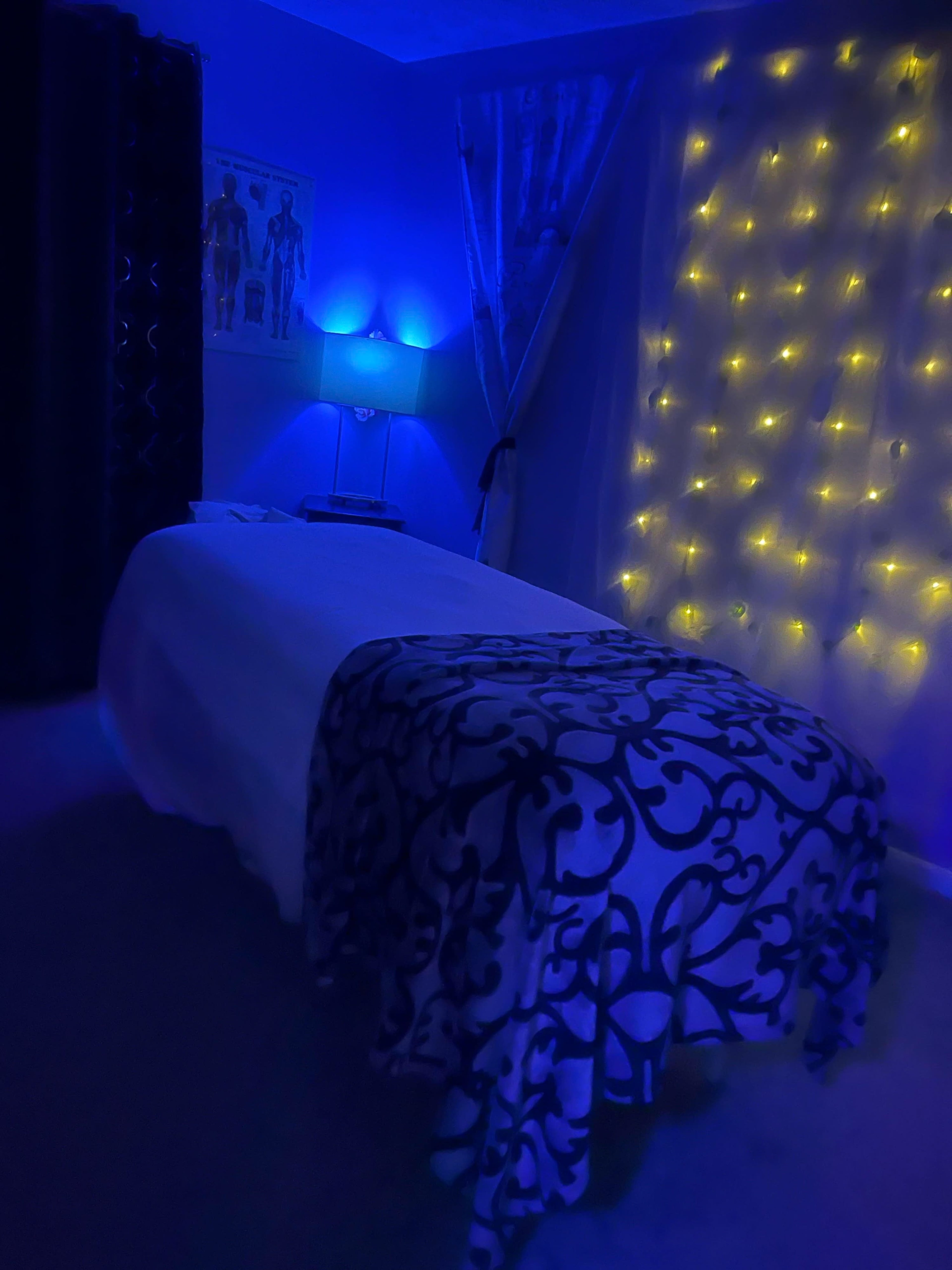 A bed with a dark patterned blanket is illuminated by blue light and surrounded by fairy lights in a dimly lit room.