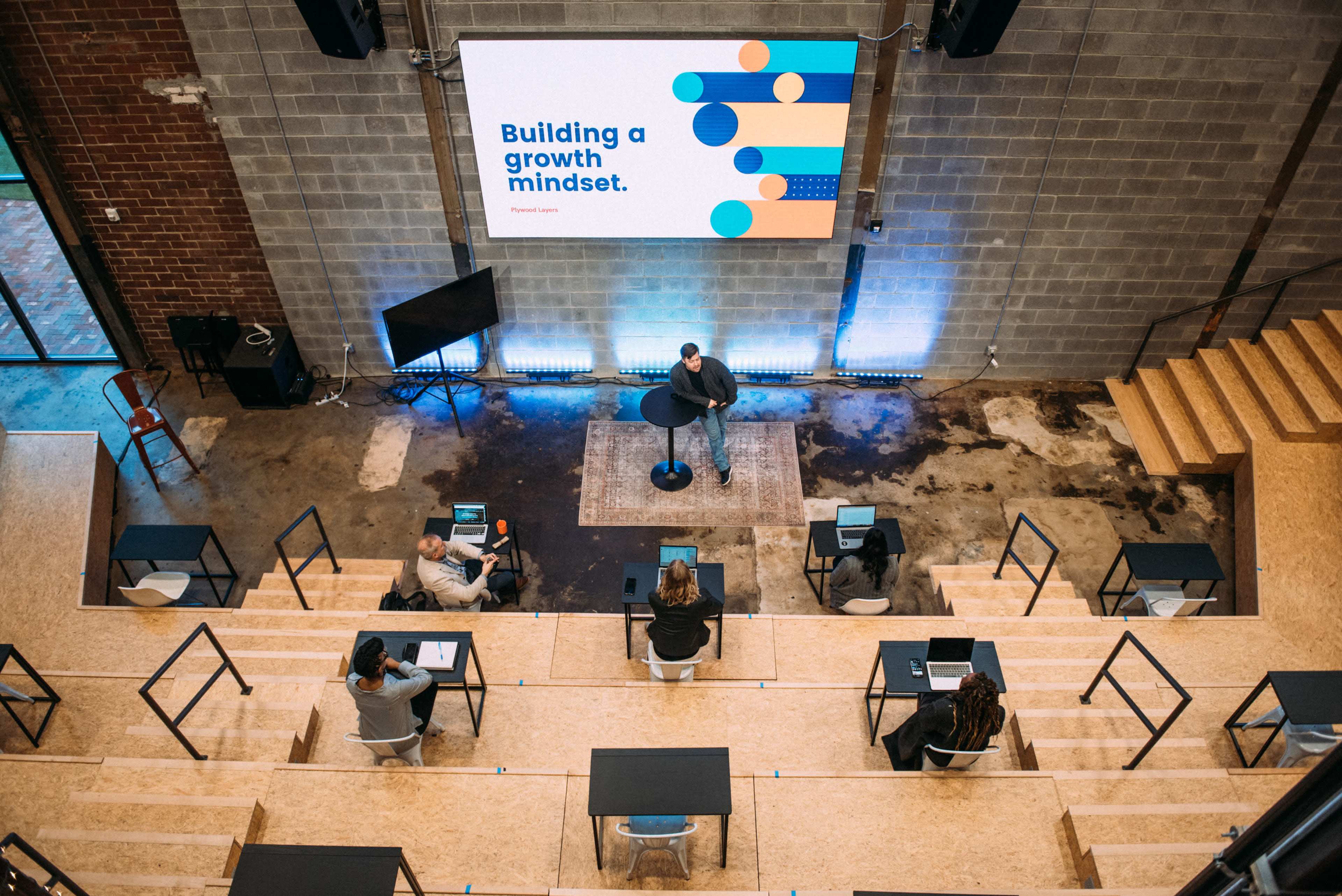 A speaker stands at a podium in front of a screen displaying "Building a growth mindset," while an audience listens attentively from tiered seating.