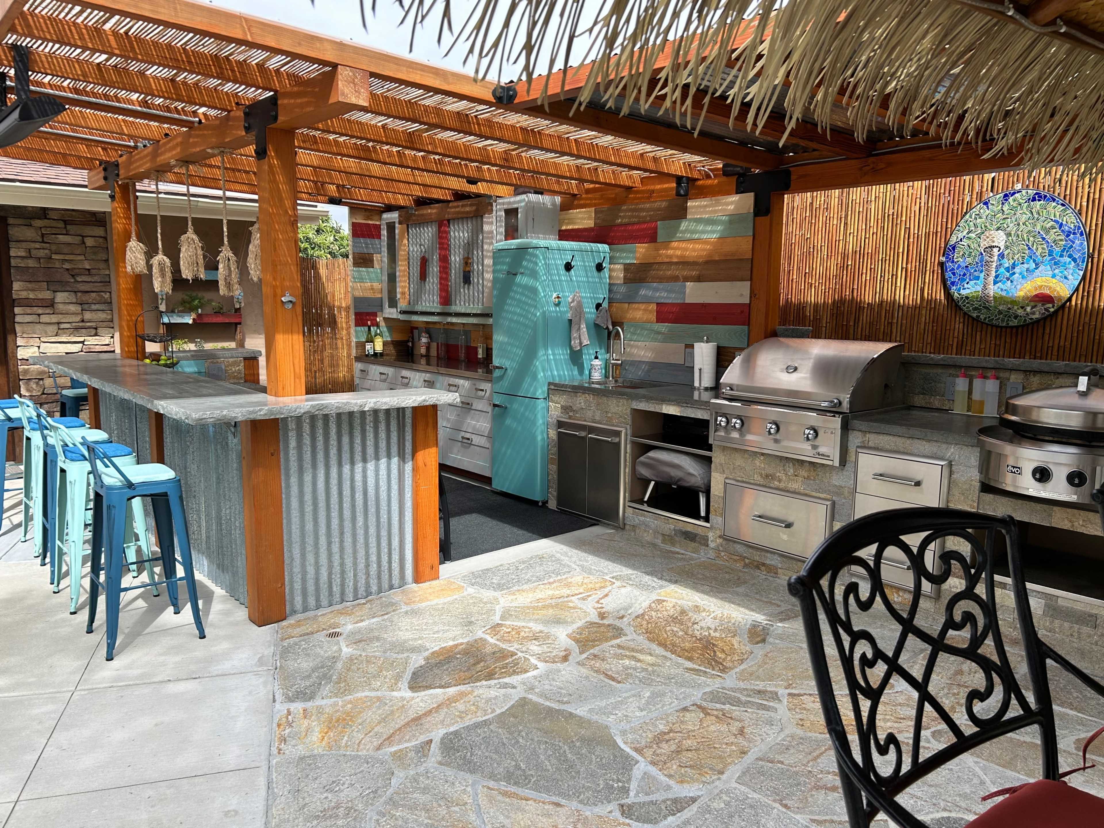 The outdoor kitchen features a turquoise vintage-style refrigerator, stainless steel appliances, and a rustic stone and metal bar area with blue stools.