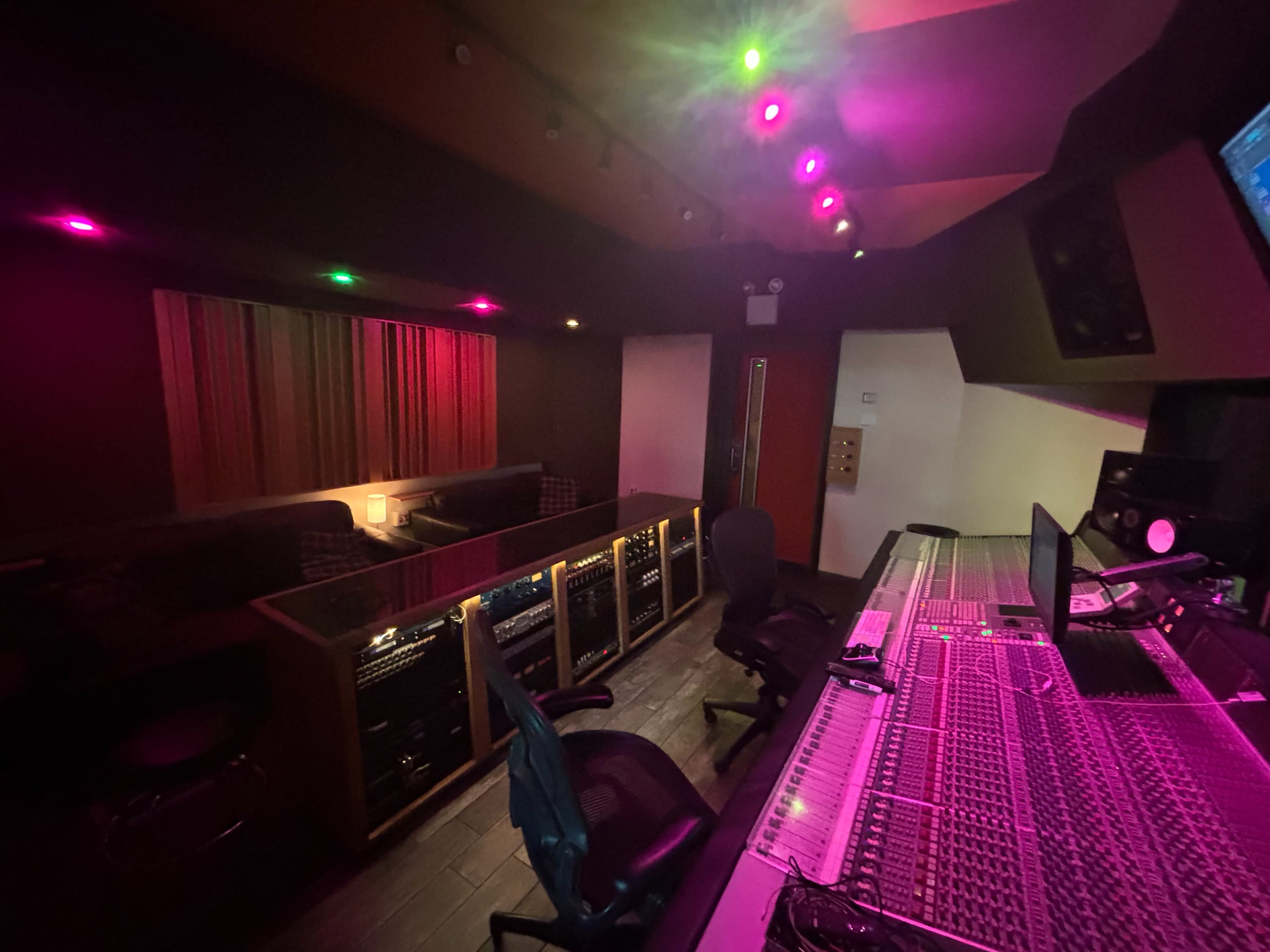 A music studio features a control desk with mixing equipment and several sound racks, illuminated by colorful lights in a dimly lit space.