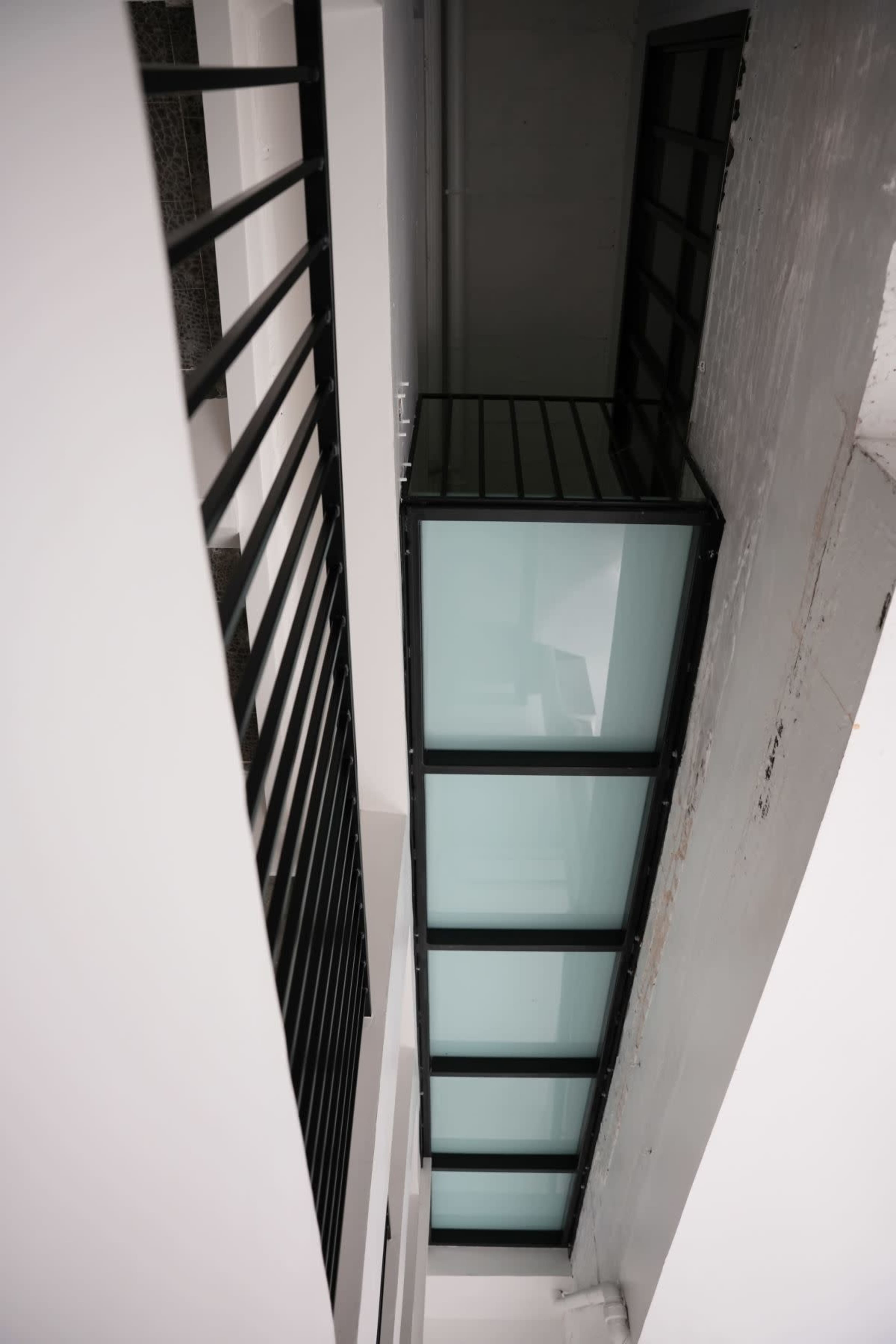The image shows a high-angle view of a narrow glass walkway connecting two sections of a building, framed by black railings and white walls.