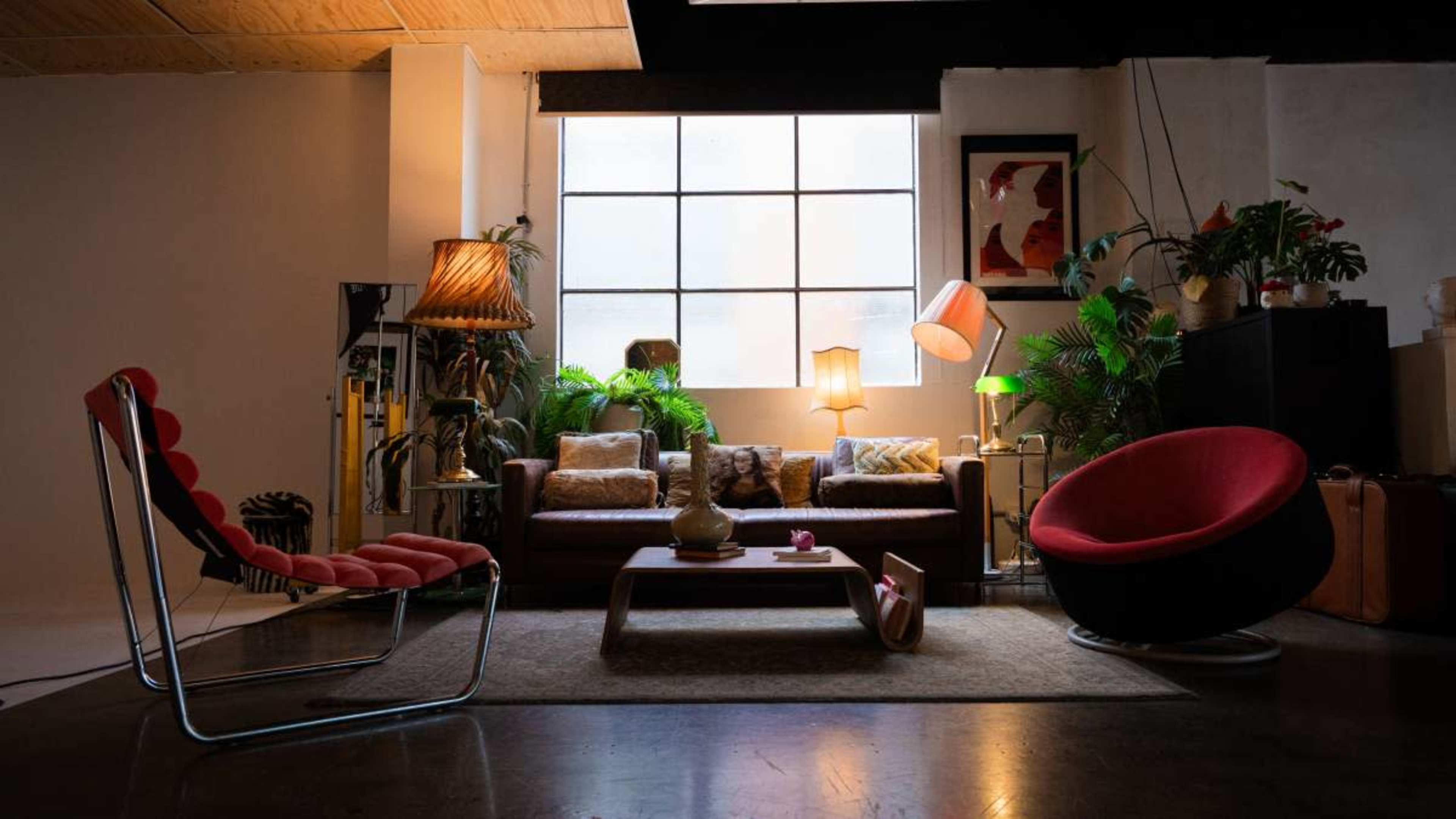 A stylish living room features a leather sofa, modern chairs, plants, and various lamps, illuminated by large windows.